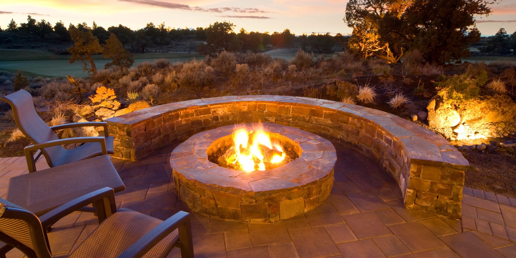 A backyard fire pit with a fire burning in it, surrounded by a stone knee wall and chairs