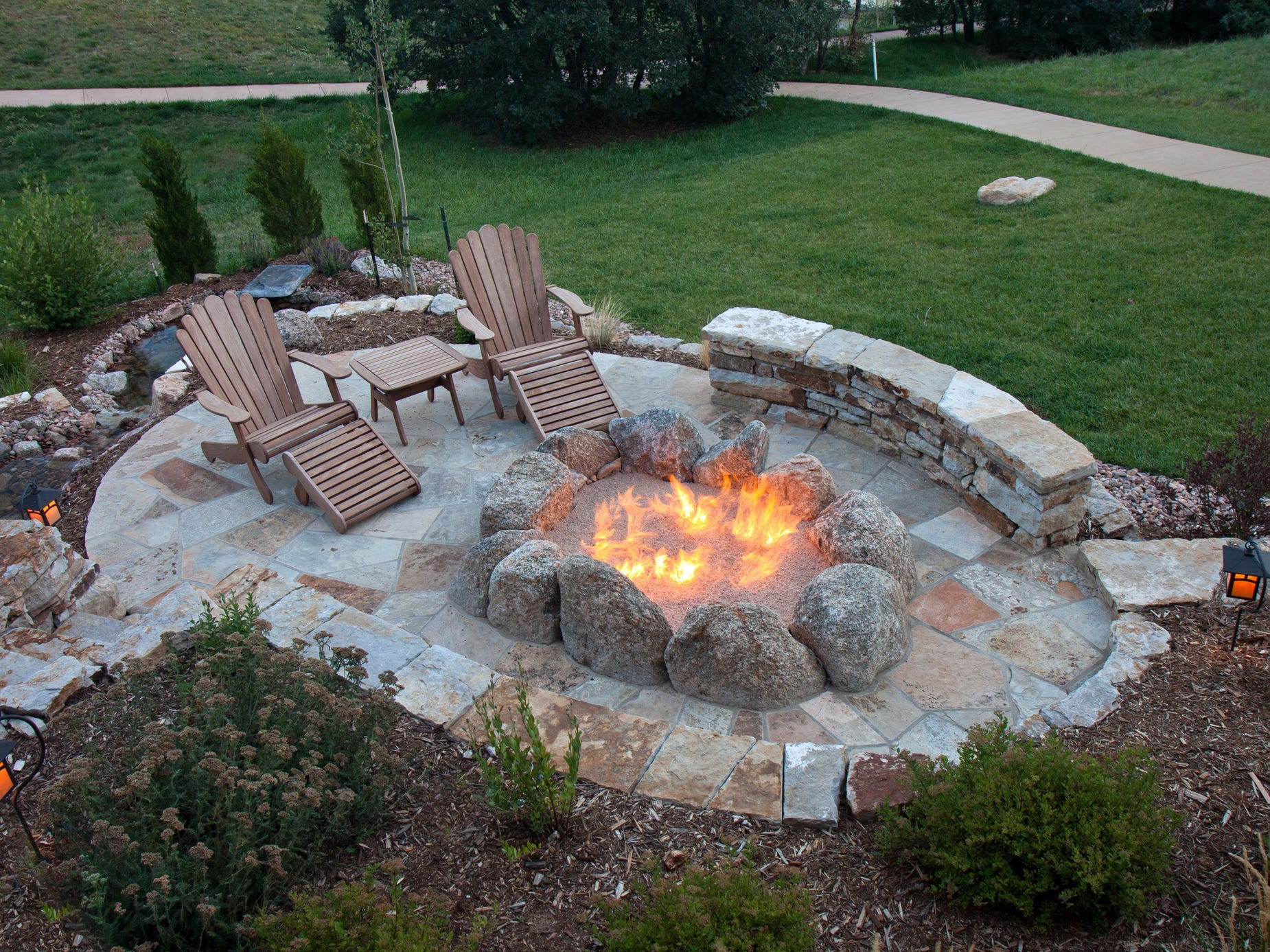 An oval fire pit area with wooden lounge chairs enclosed by a stone wall