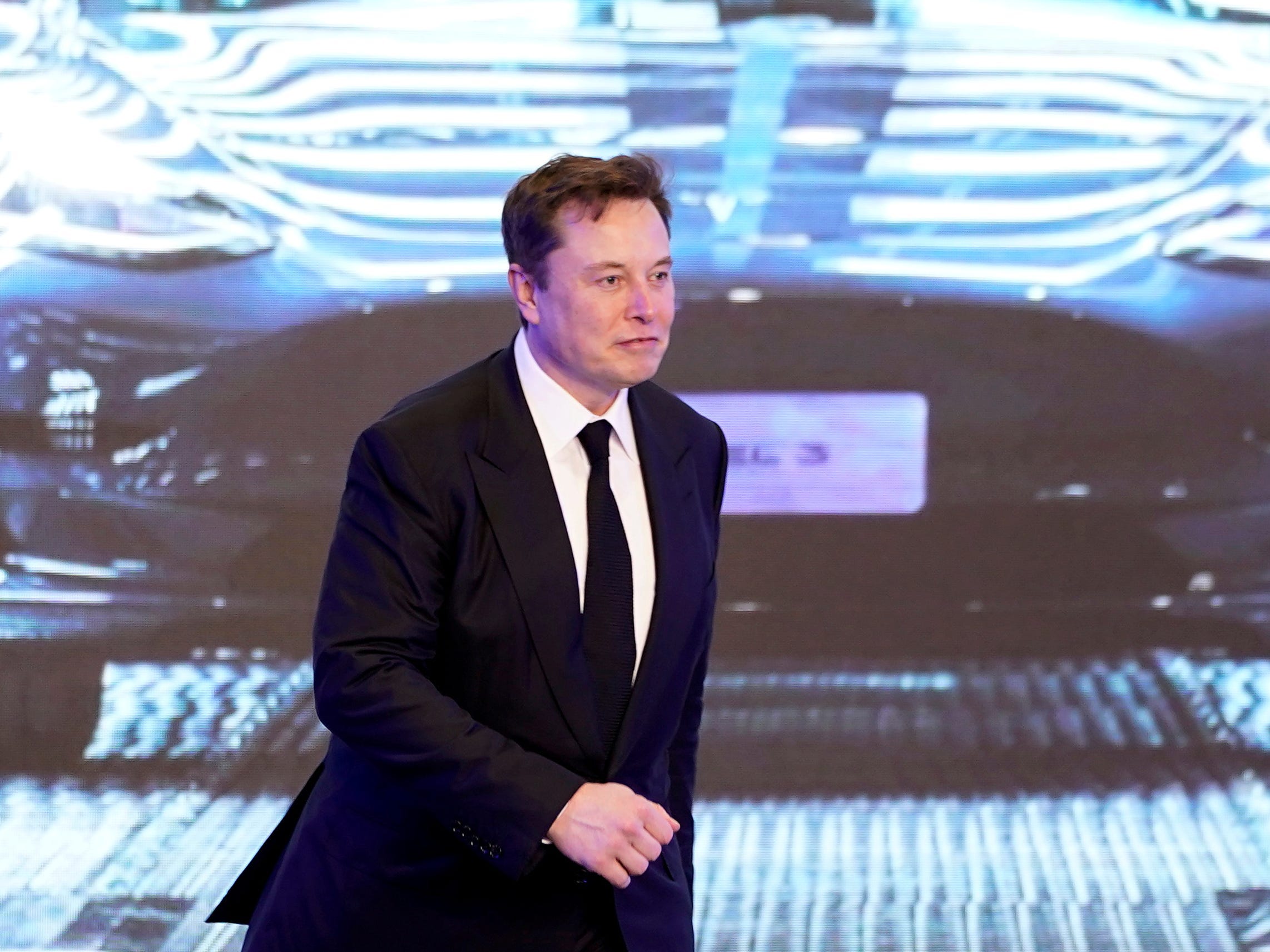 Tesla CEO Elon Musk in a black suit walks on stage in front of an image of a Model Y vehicle