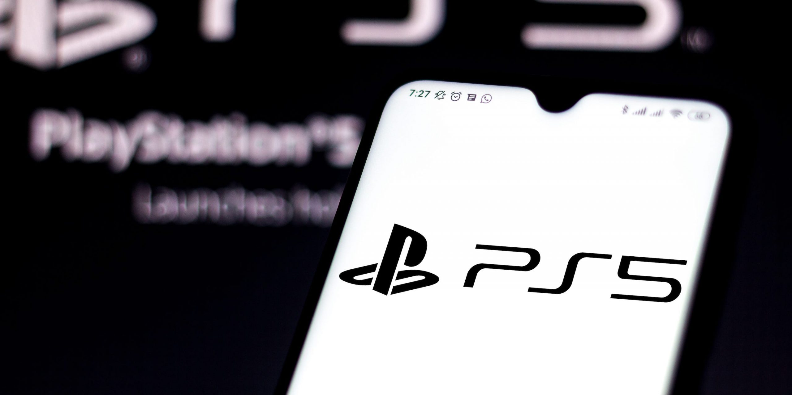 A smartphone displaying the PS5 logo. Behind it, the PS5 logo is written larger in white on a black screen.