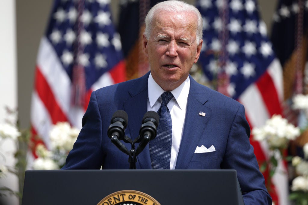 President Joe Biden wears a blue suit jacket and tie while giving a speech in front of two American flags.