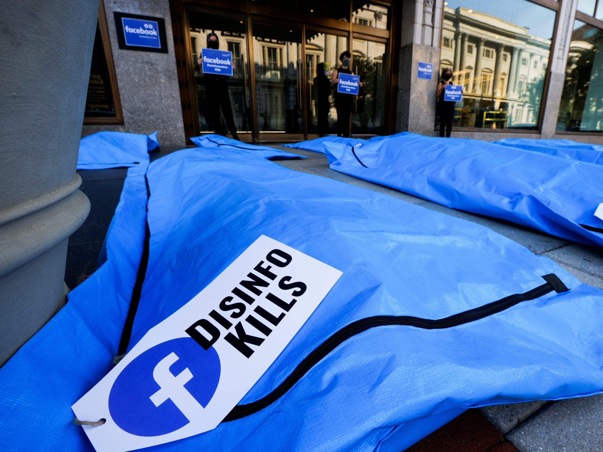 a blue body bag in the foreground with a tag including the facebook logo and the words "disinfo kills"