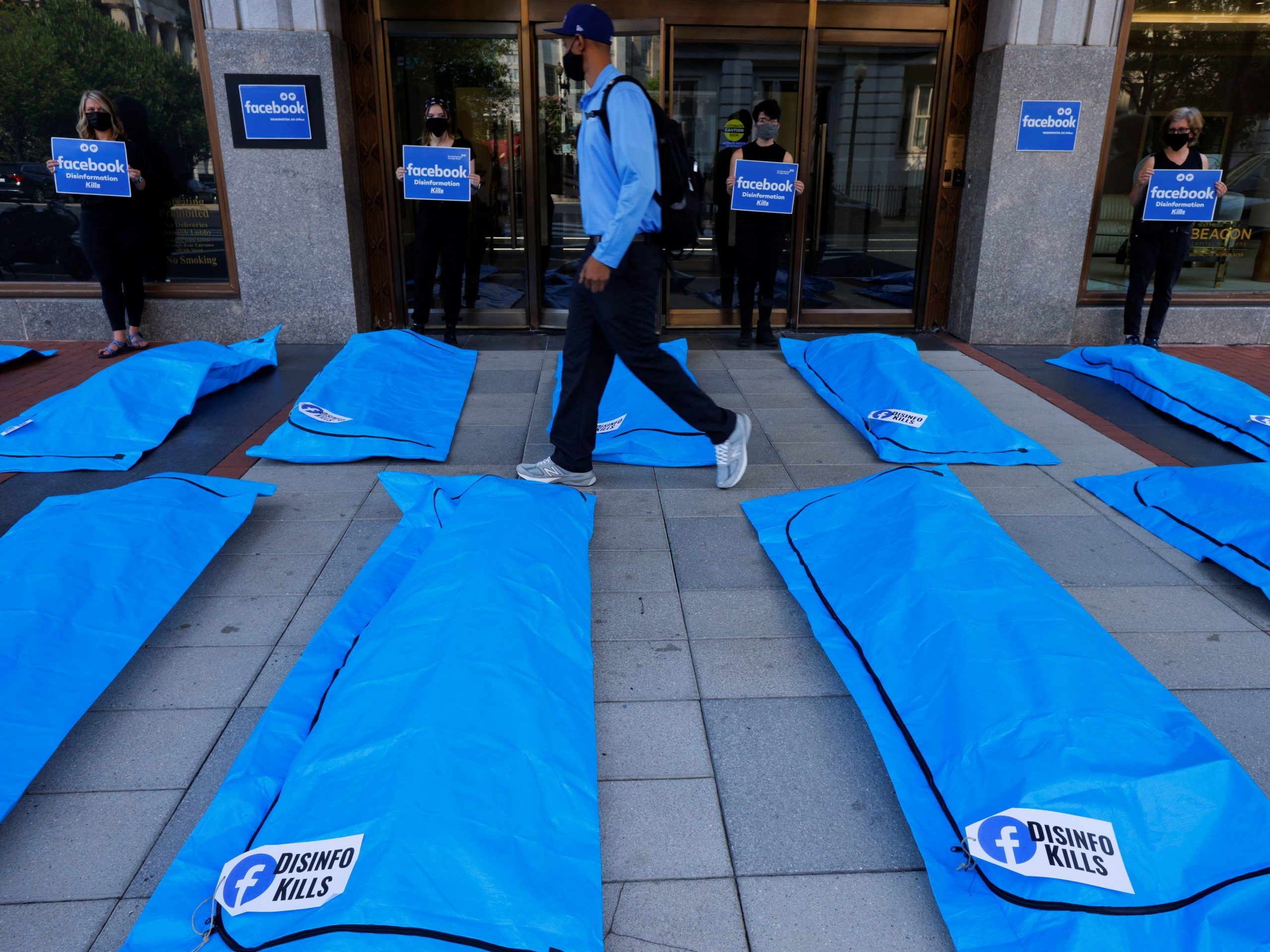 a pedestrian walks through a protest between two rows of blue body bags that read "disinfo kills." the bags are surrounded by activists holding signs that read "facebook disinformation kills"