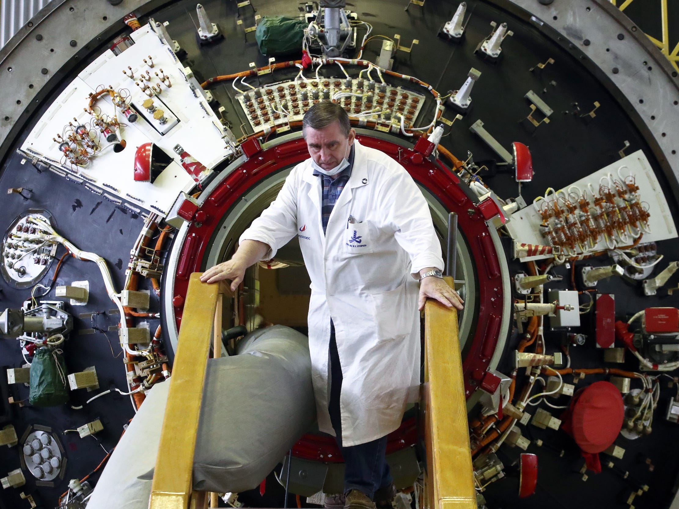 man in white lab coat stands in front of nauka module port opening in lab room