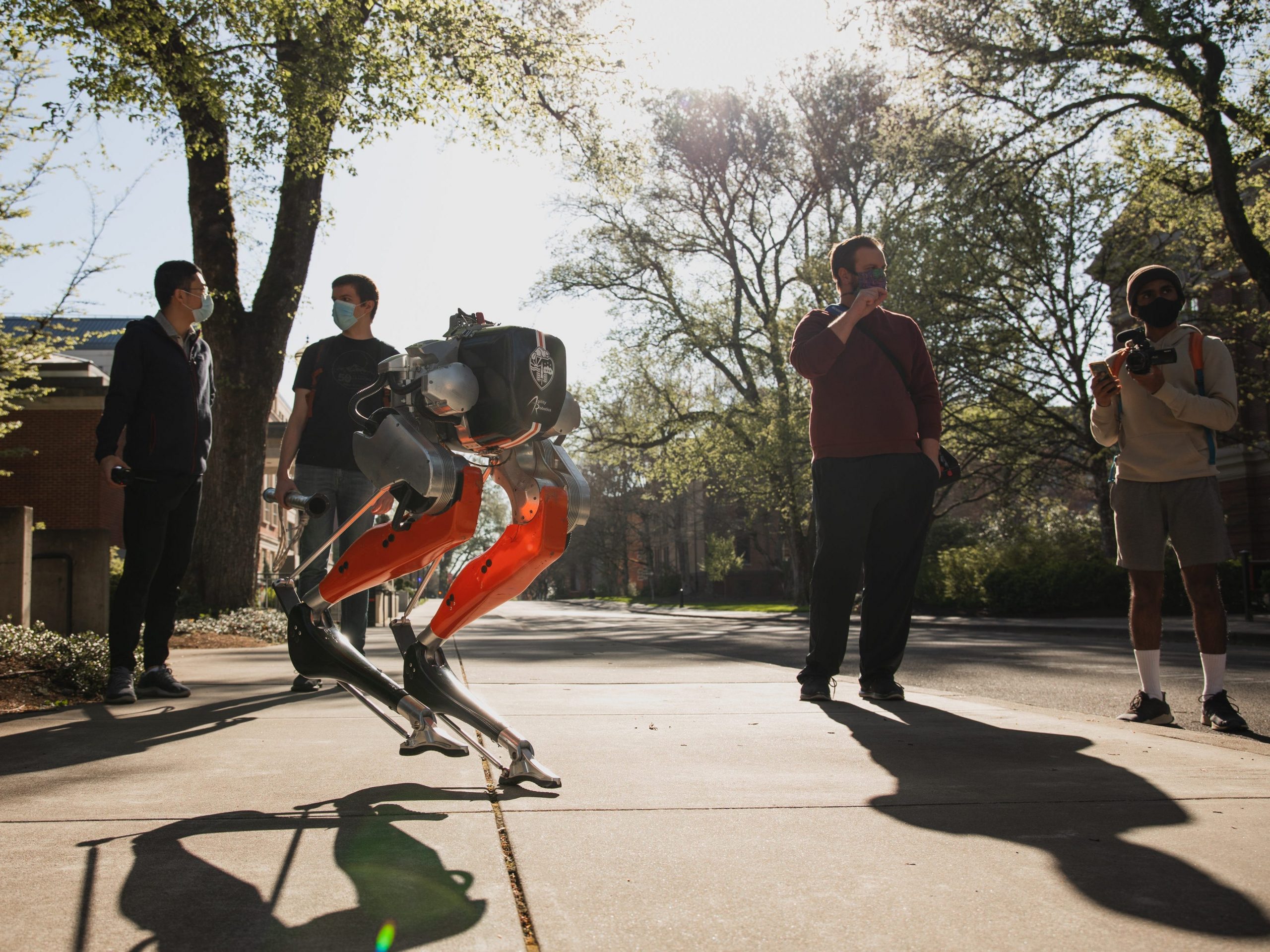 a bipedal robot named Cassie running a 5K outdoors with trees and four people in the background