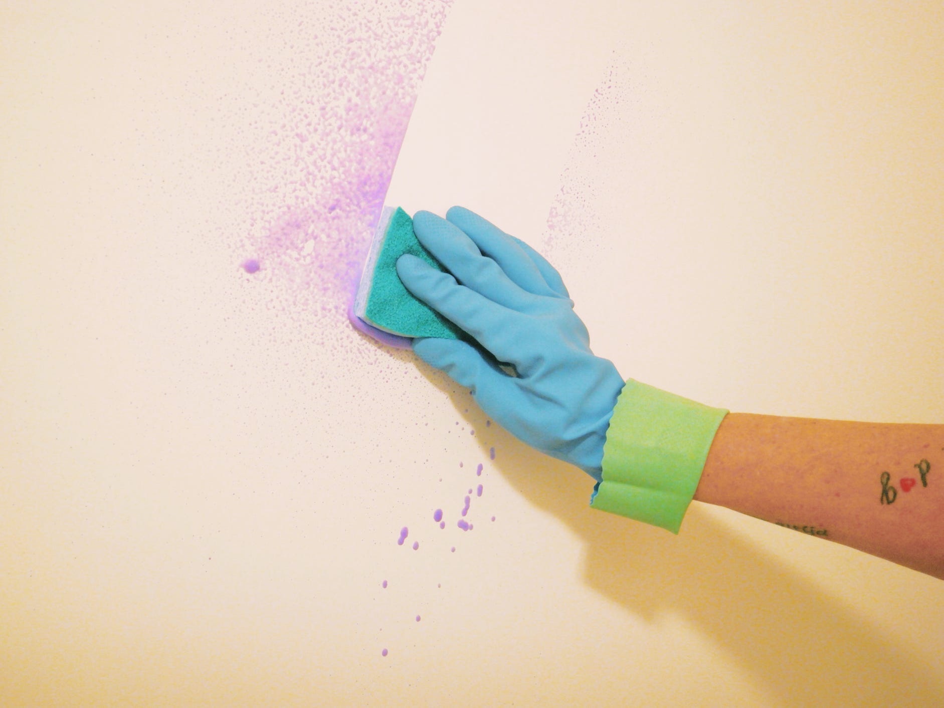 A gloved hang wiping purple cleaning spray off the wall with a sponge