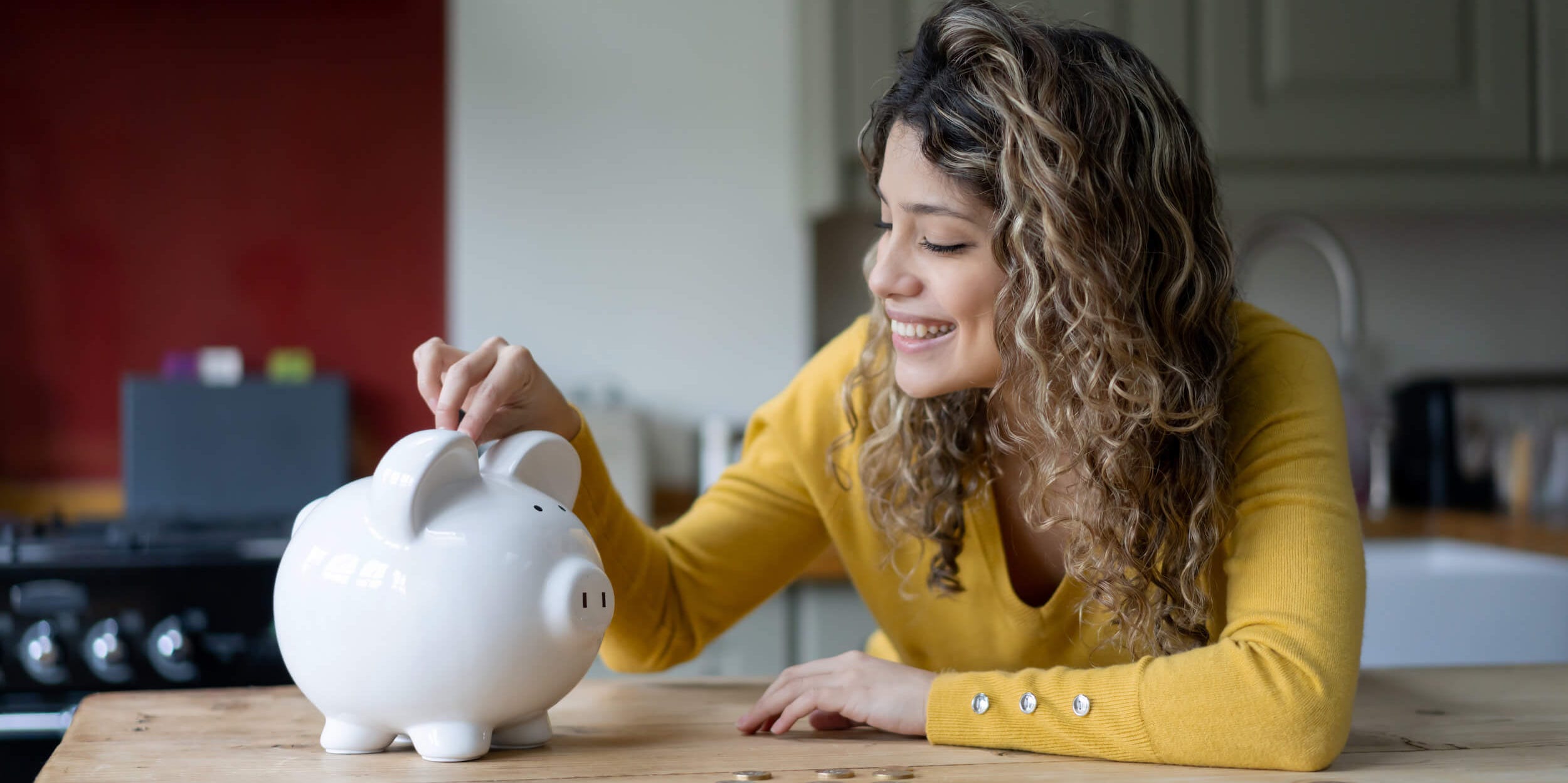 A cheerful young woman with curly hair at home saving coins into her piggy bank.