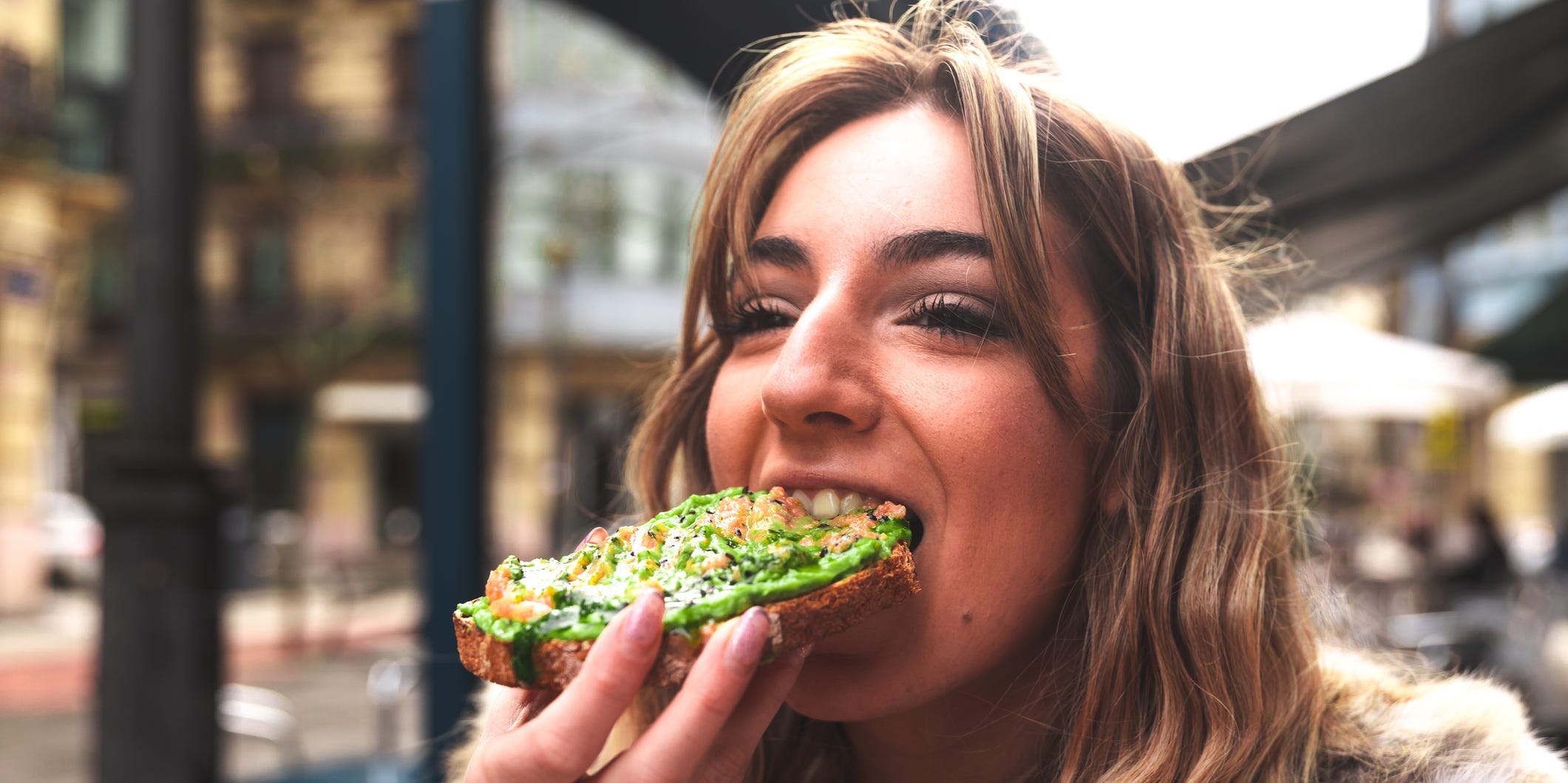 A young woman bites into a slice of avocado toast while standing outside.