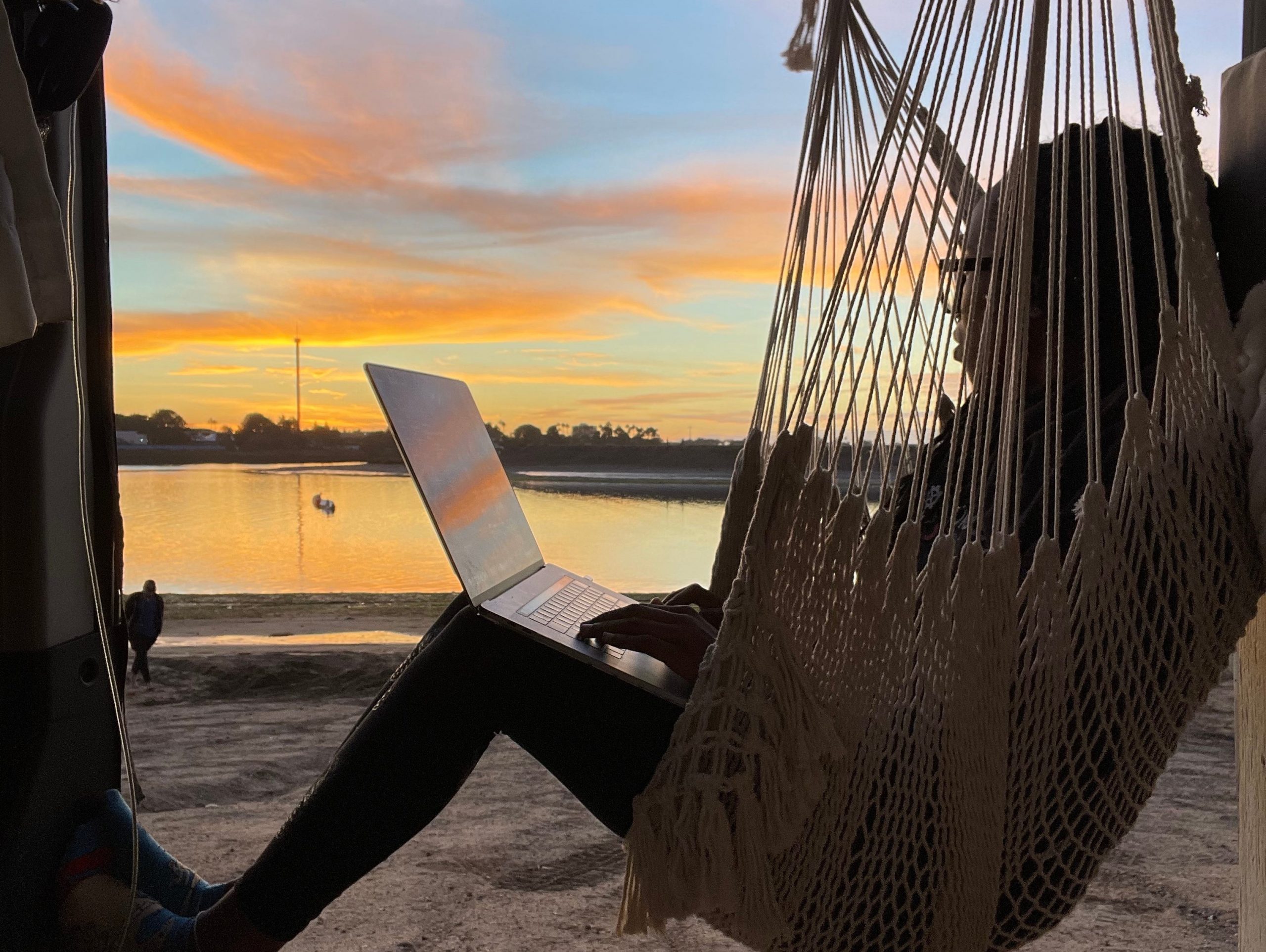 woman sitting in a hammock holding a laptop next to a sunset