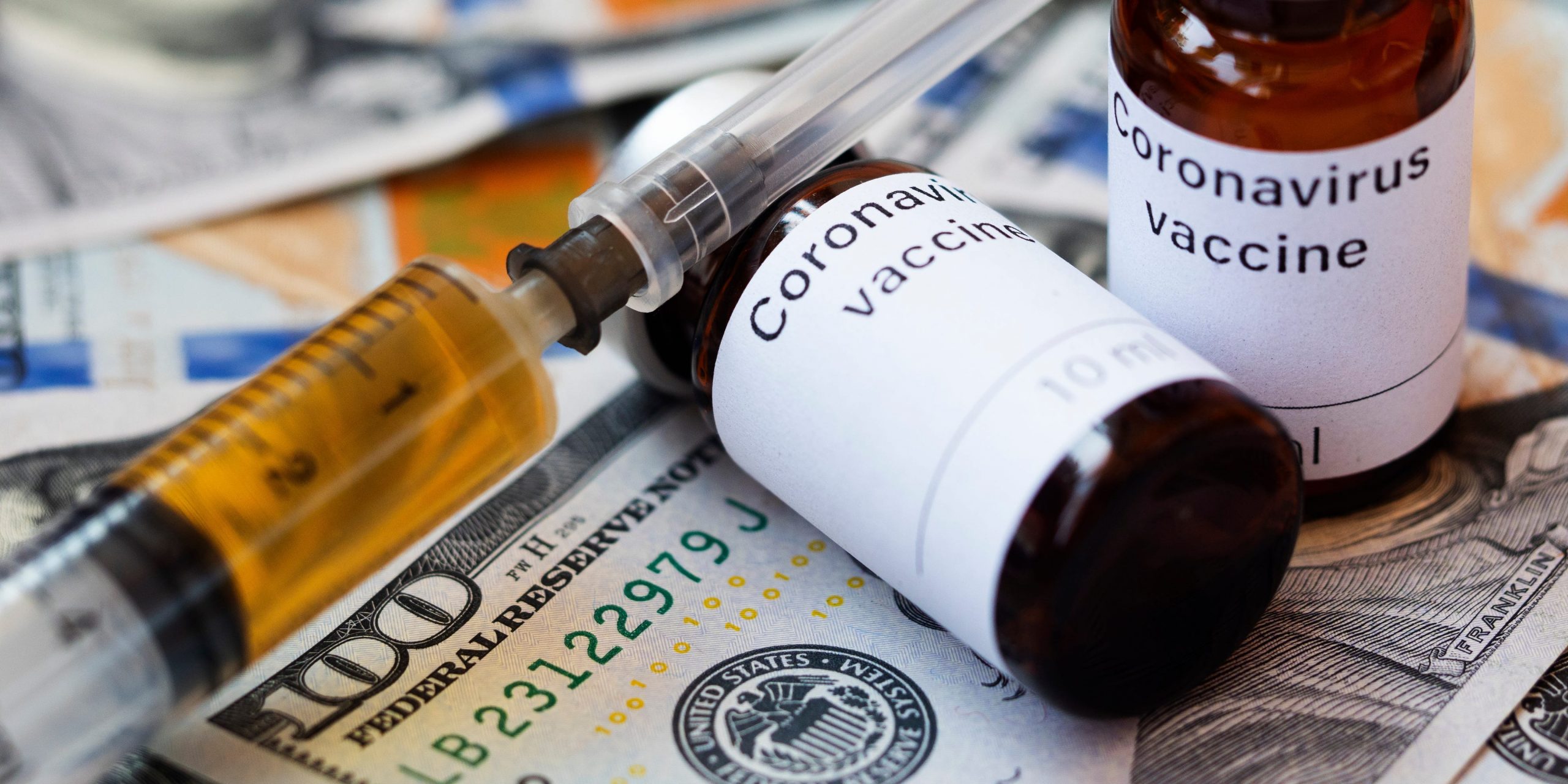 Two vials labelled Coronavirus vaccine and a syringe seen displayed on one hundred US dollar banknotes.