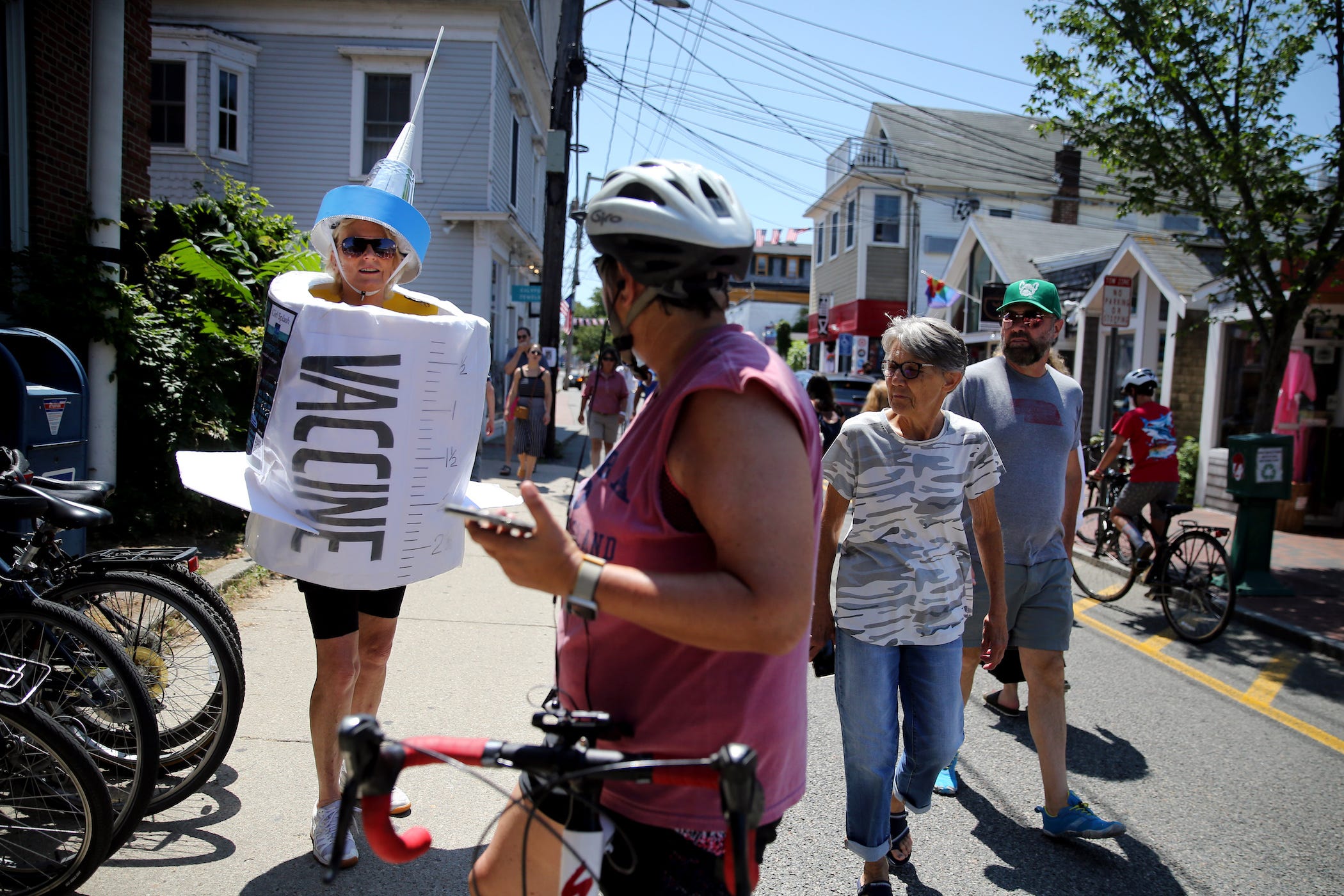 woman wearing vaccine costume walks down street with bikers, pedestrians watching