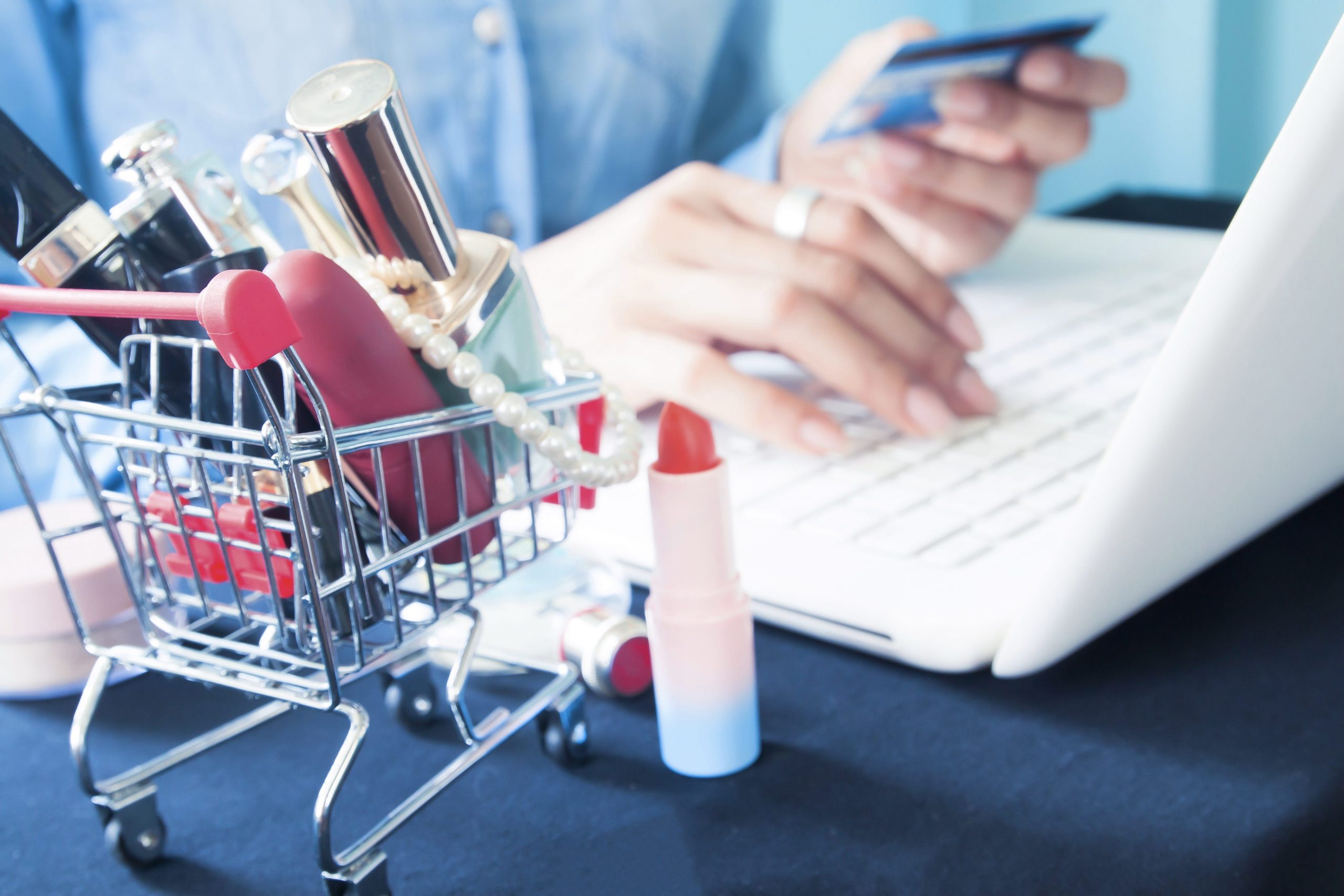 A mini shopping cart with makeup products next to a person entering their credit card information on a laptop