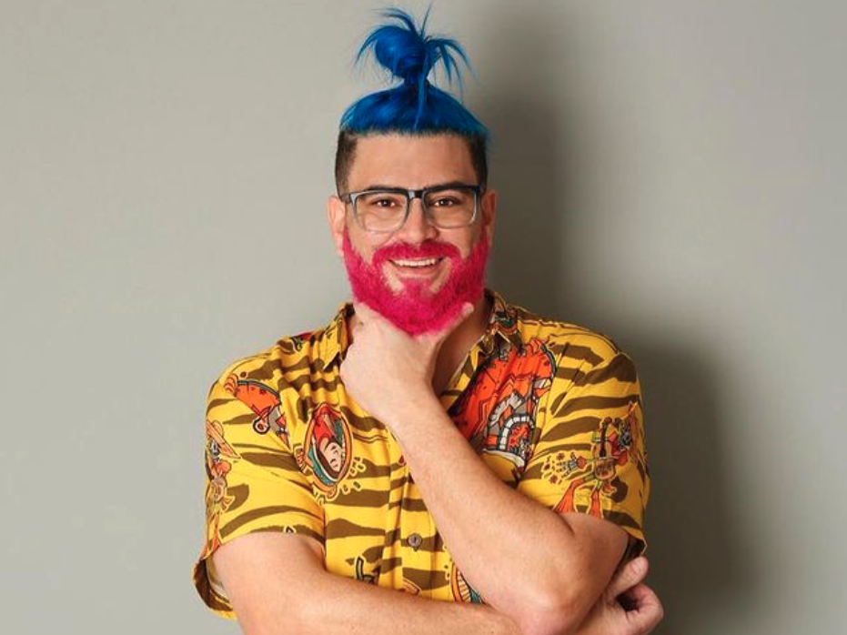 Raulino de Oliveira Maciel, the Brazilian streamer, in a yellow shift with a pink beard and blue hair tied up in a bun. He is similing and has his hand resting under his chin.