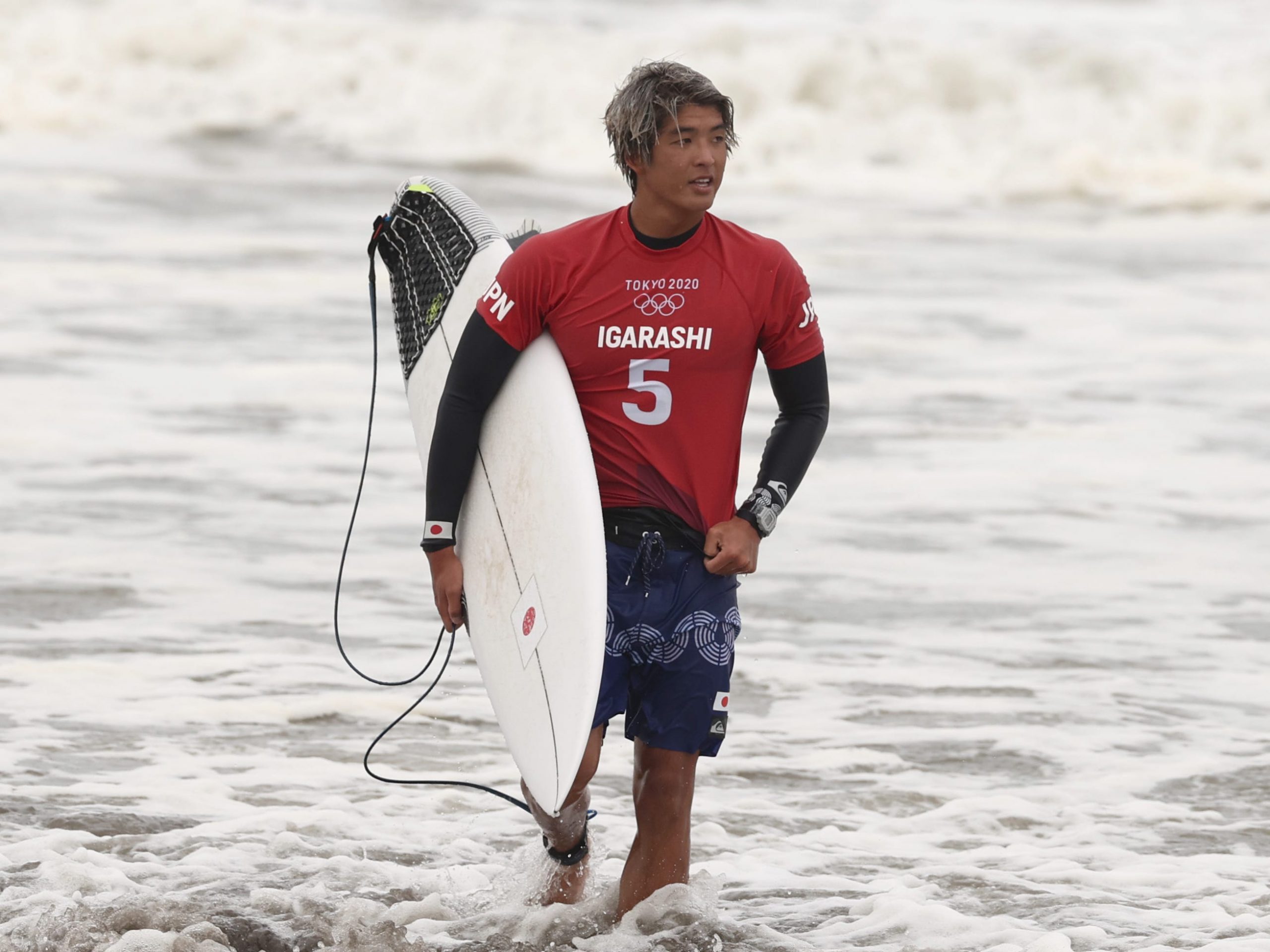 surfer kanoa igarashi stepping out of the surf holding a white surfboard and wearing a red team japan shirt