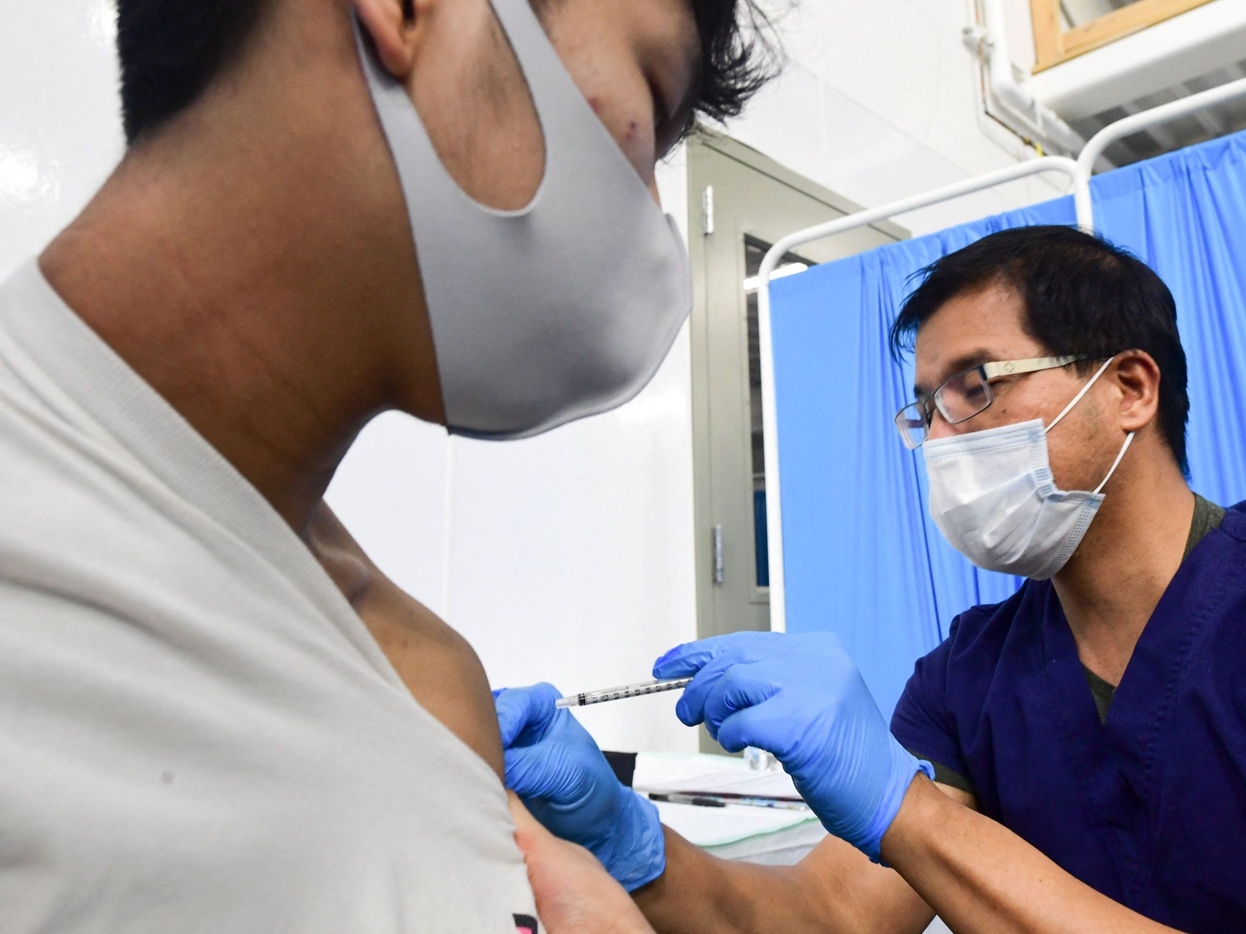 Registered Nurse Robert Orallo, wearing a face mask and blue gloves, injects a syringe containing a COVID-19 vaccine into a patient's arm