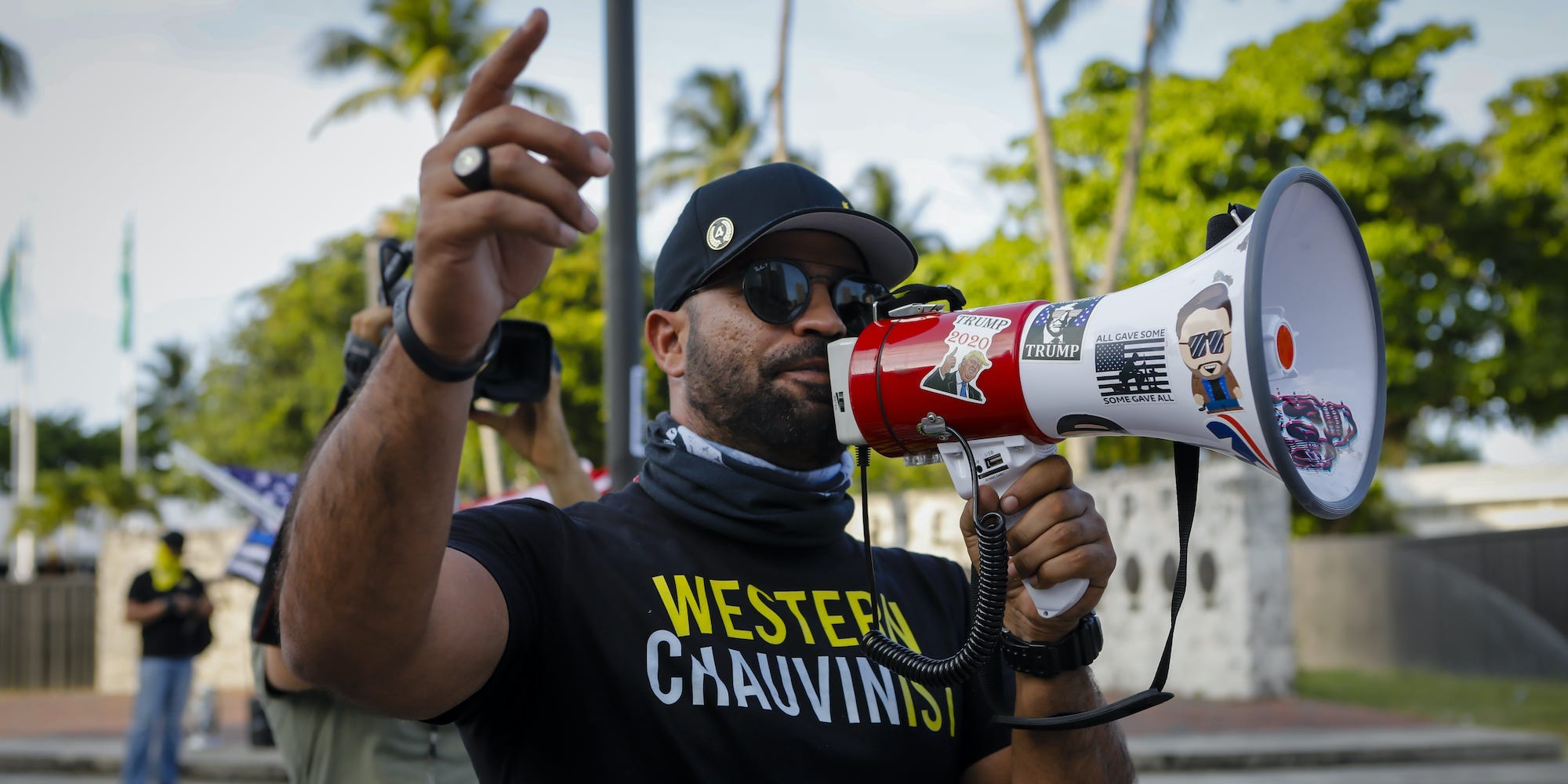 The Proud Boys leader Enrique Tarrio speaks into a megaphone, wearing a black shirt, sunglasses and a black shirt that reads "Western Chauvinist."