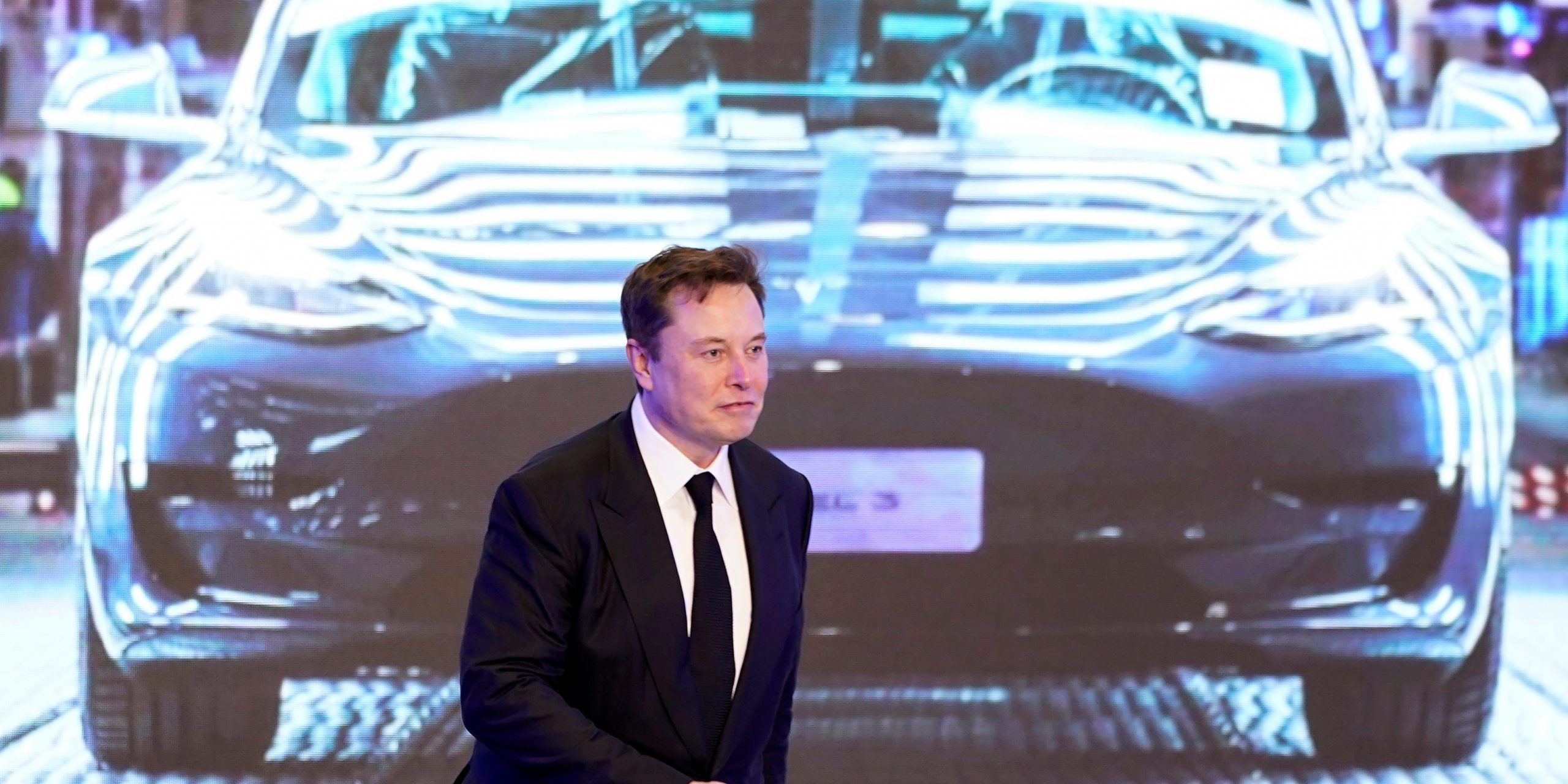 Tesla CEO Elon Musk in a black suit walks on stage in front of an image of a Model Y vehicle