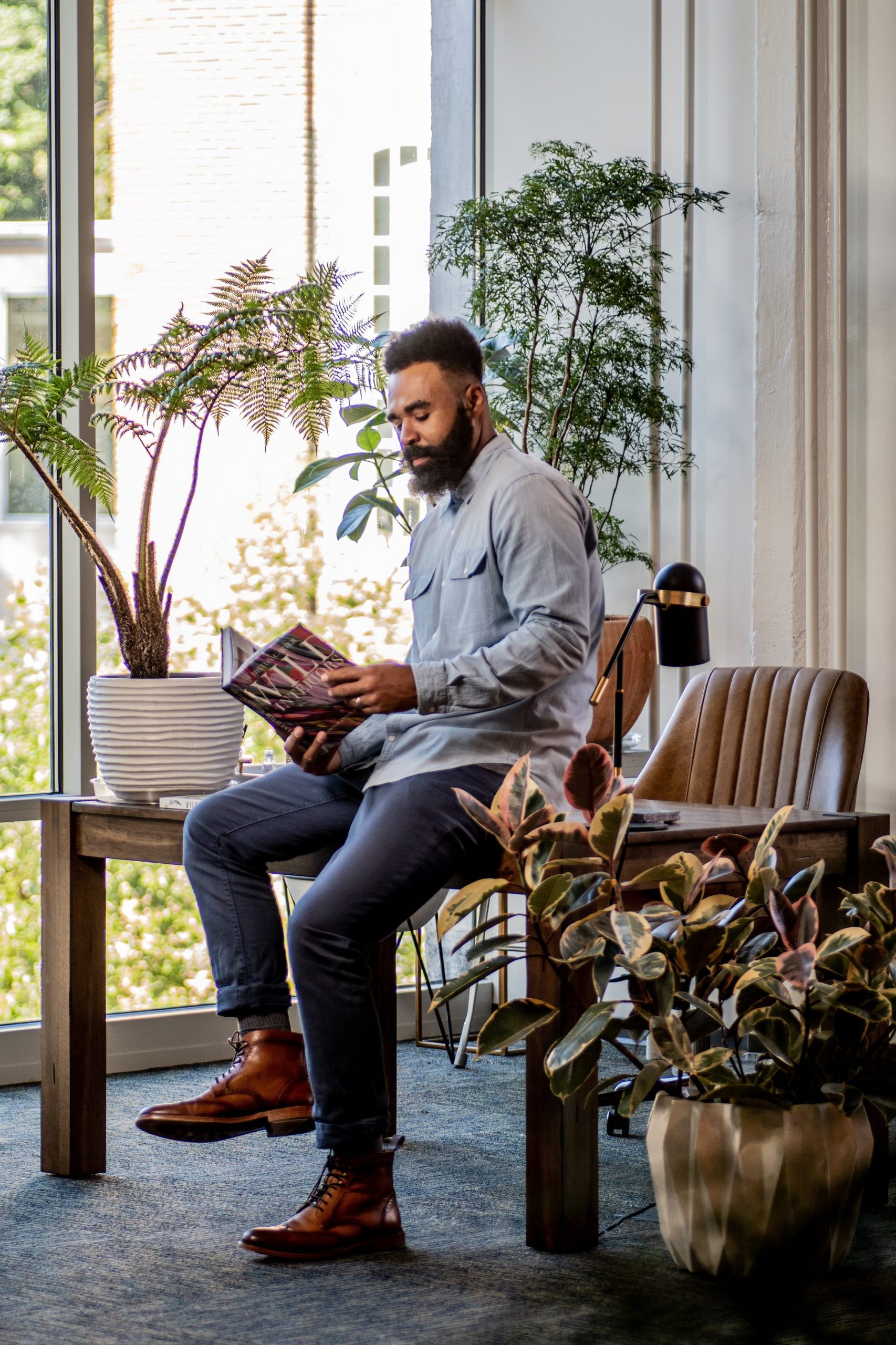 Hilton Carter sits on a desk and reads a book in a room filled with plants.