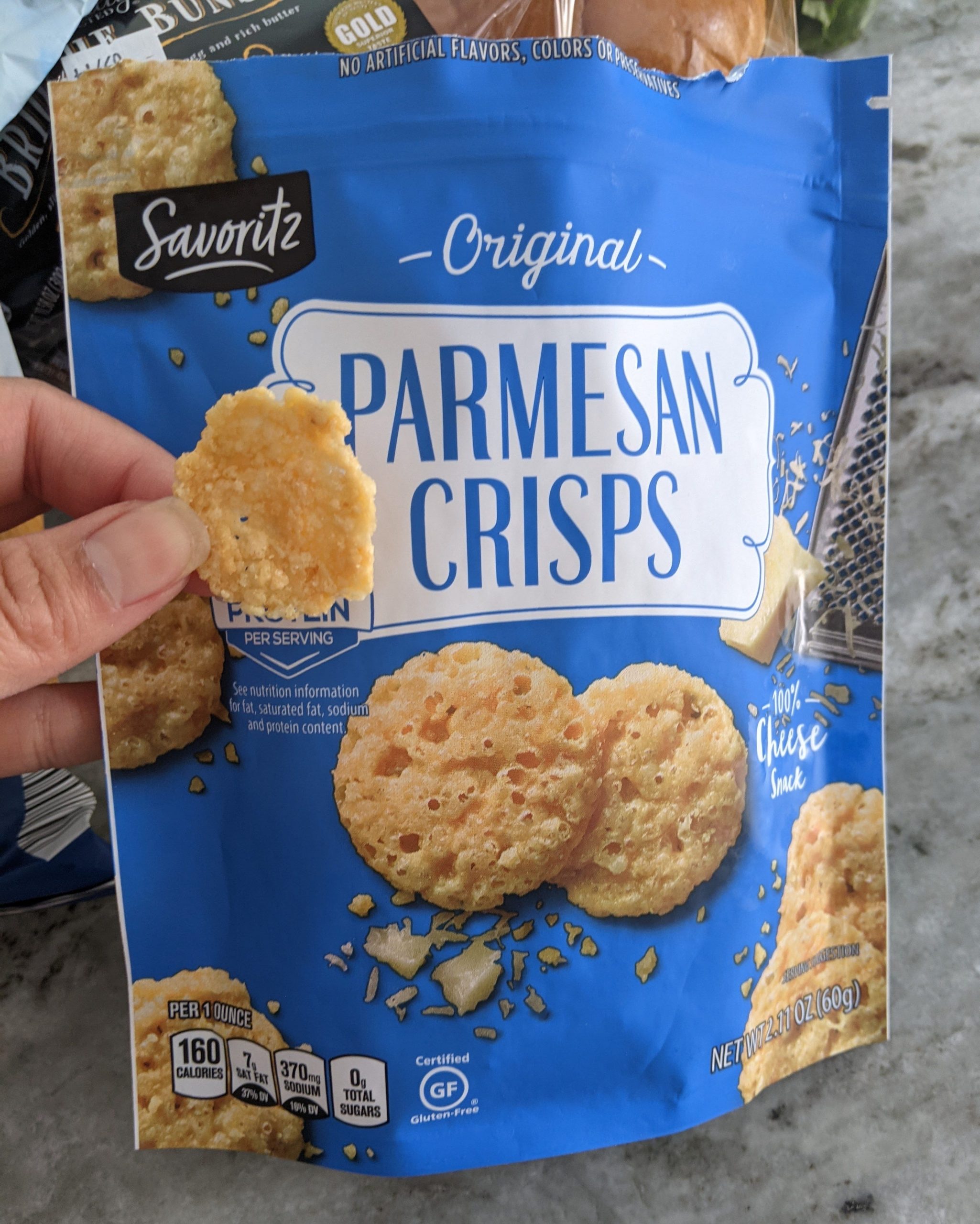 A blue bag of Aldi's Parmesan crisps and a hand holding one of the crisps in front