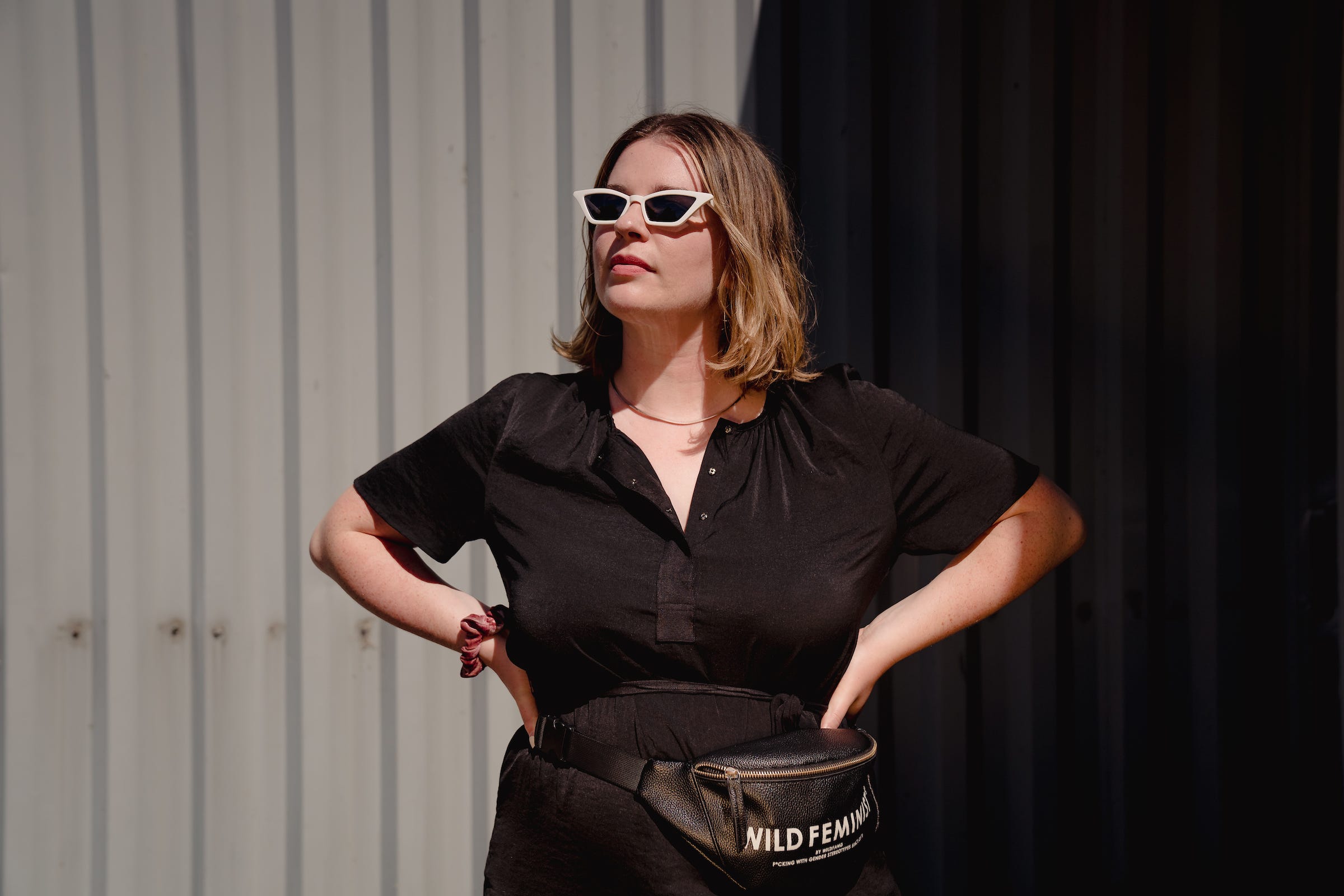 Tori Dunlap wearing a black short-sleeve dress with a necklace, a pair of white sunglasses, and a fanny pack that says "Wild Feminist."