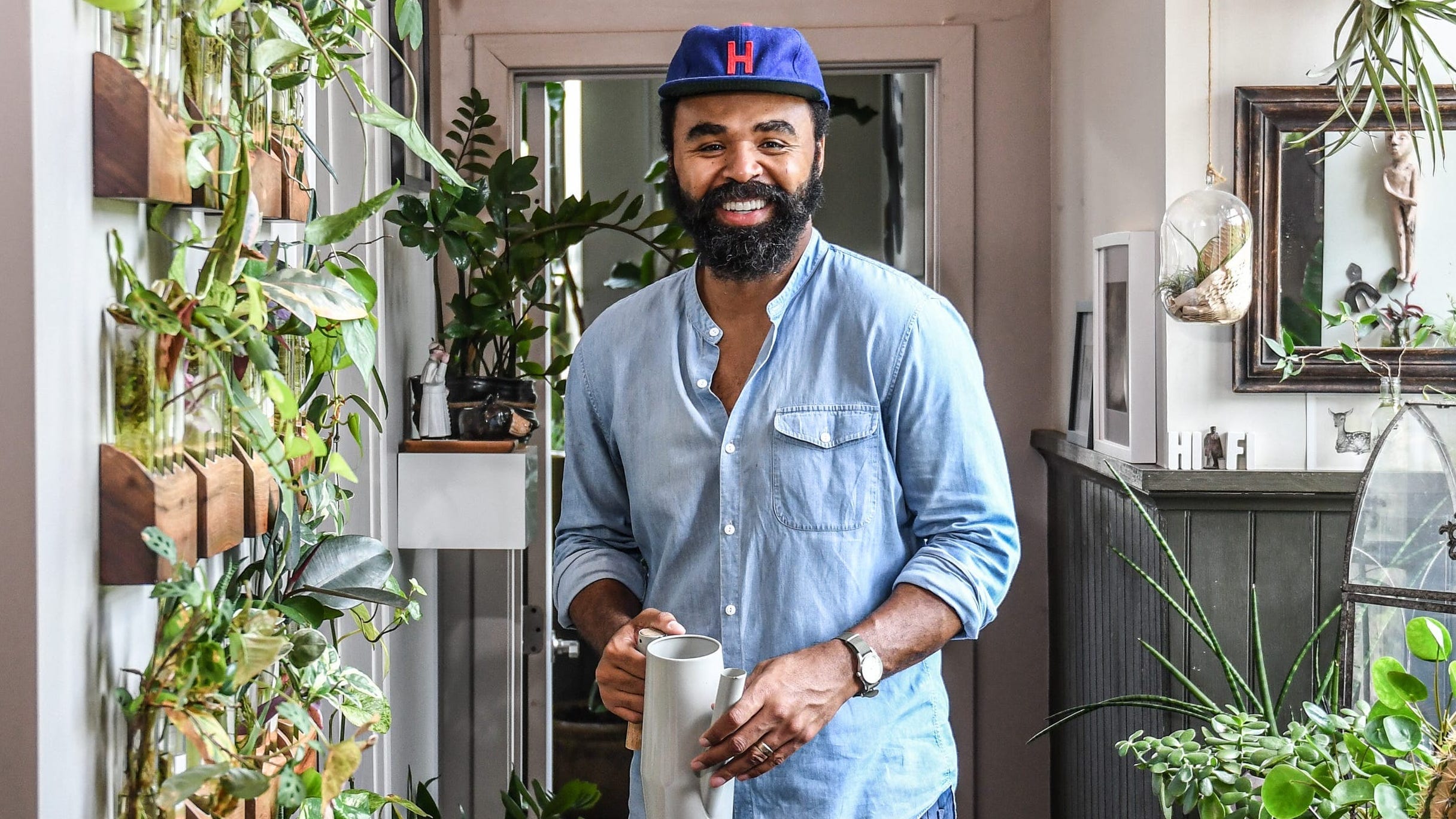 Hilton Carter smiling and posing in a hallway filled with plants, wall hangings, and string lights.