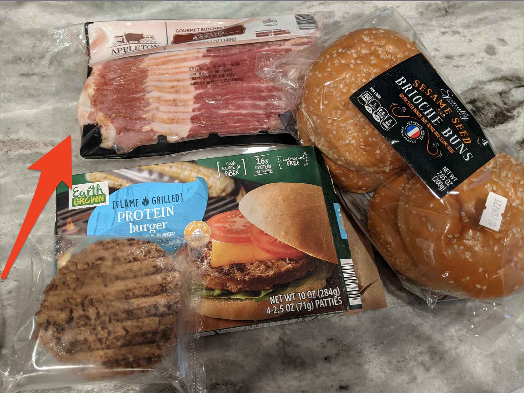 Aldi bacon, burgers, and buns on a granite counter, with a red arrow pointing to the bacon