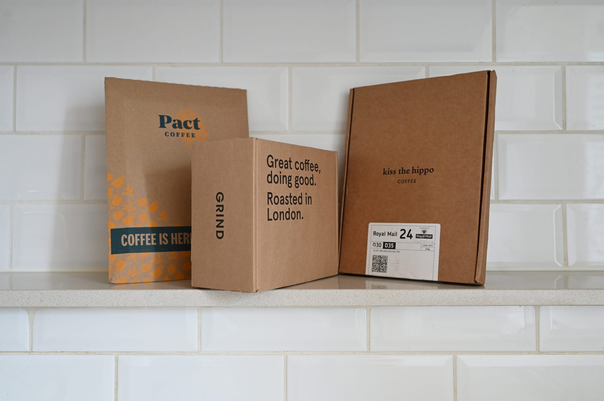 Coffee packages from Pact, Grind, and Kiss the Hippo in front of white tiles