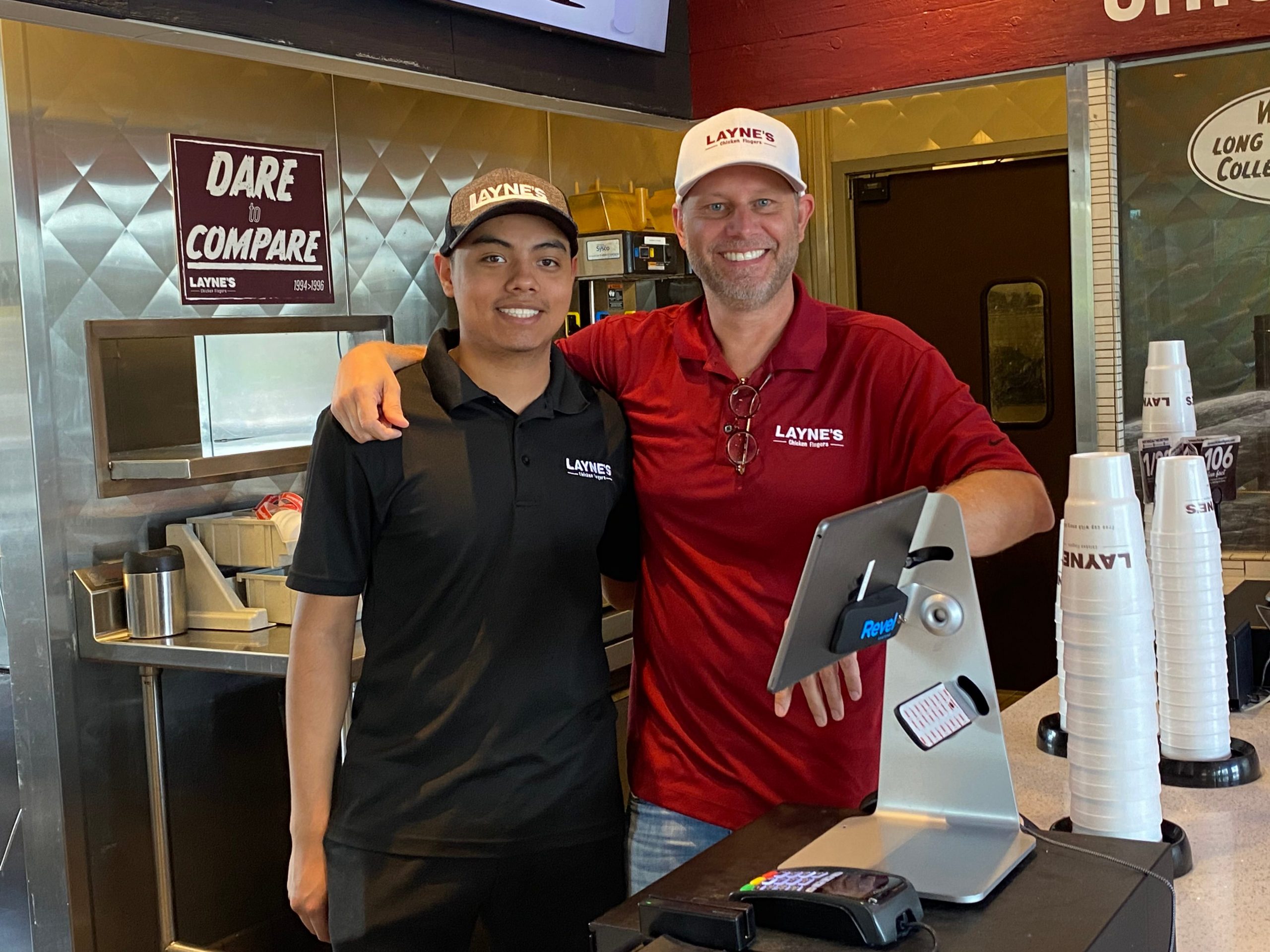 Two men stand behind the counter at a fast food restaurant smiling at the camera with their arms around each other.