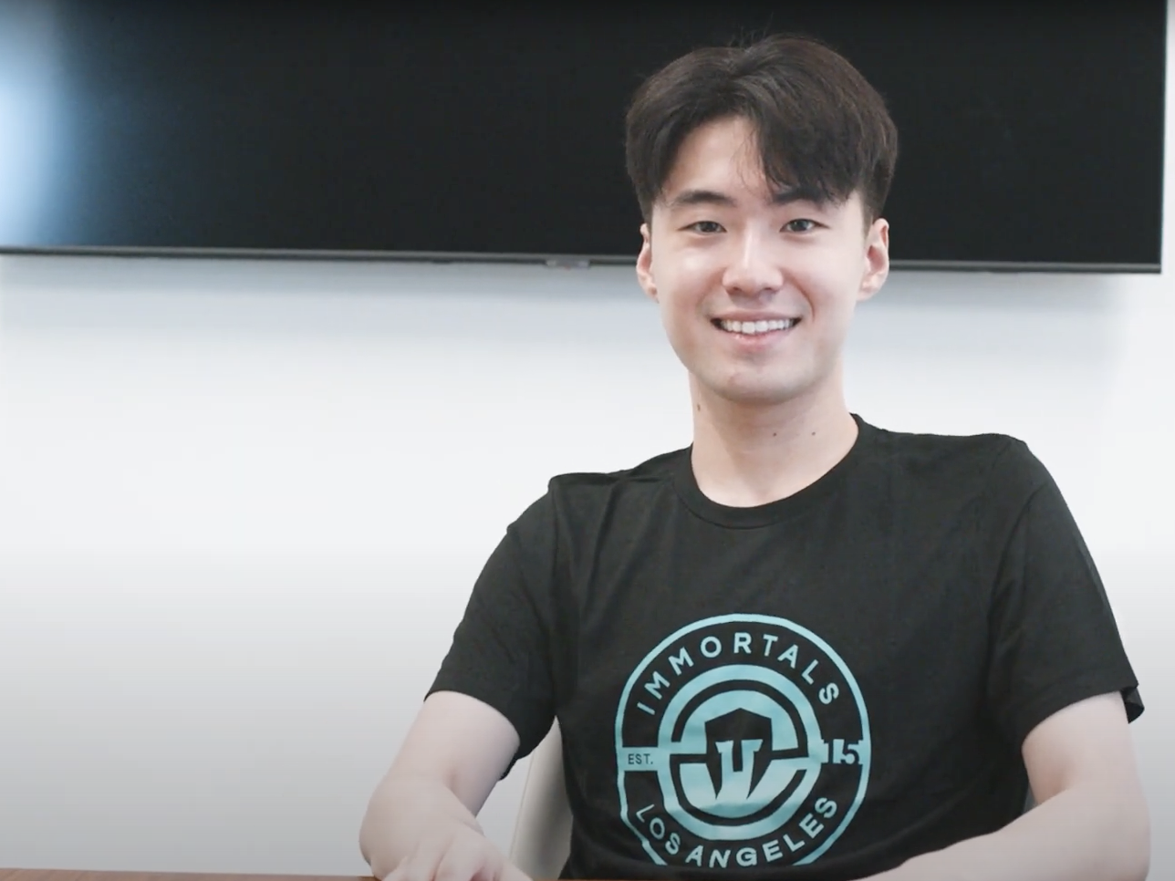 SeongHun Jang smiling in a black shirt with the Immortals Logo. He's sitting in front of a turned-off TV mounted on a white wall