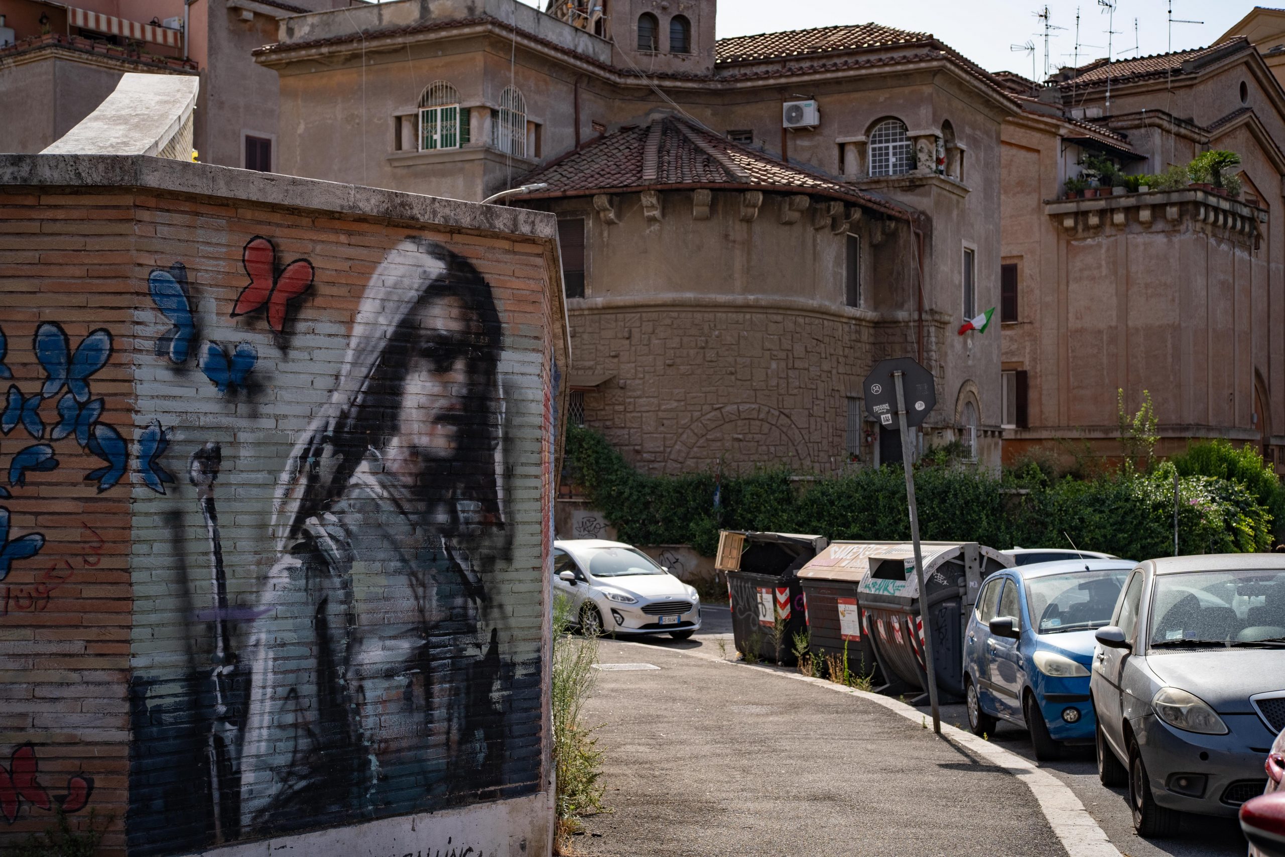 A mural is seen on a wall along a steep hilly road, with a baroque building in the background.