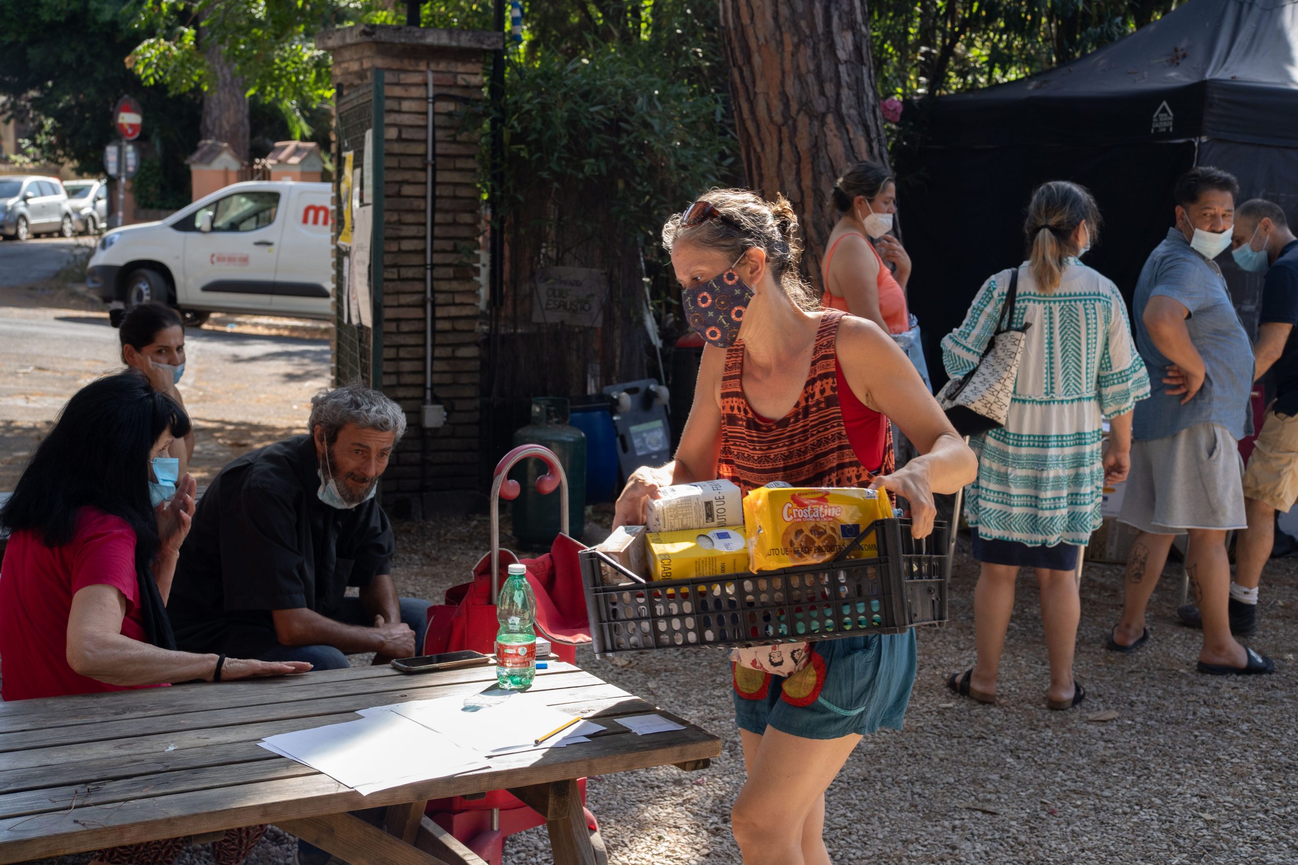 A woman is seen holding a crate of goods while others sit at tables or wait in line.