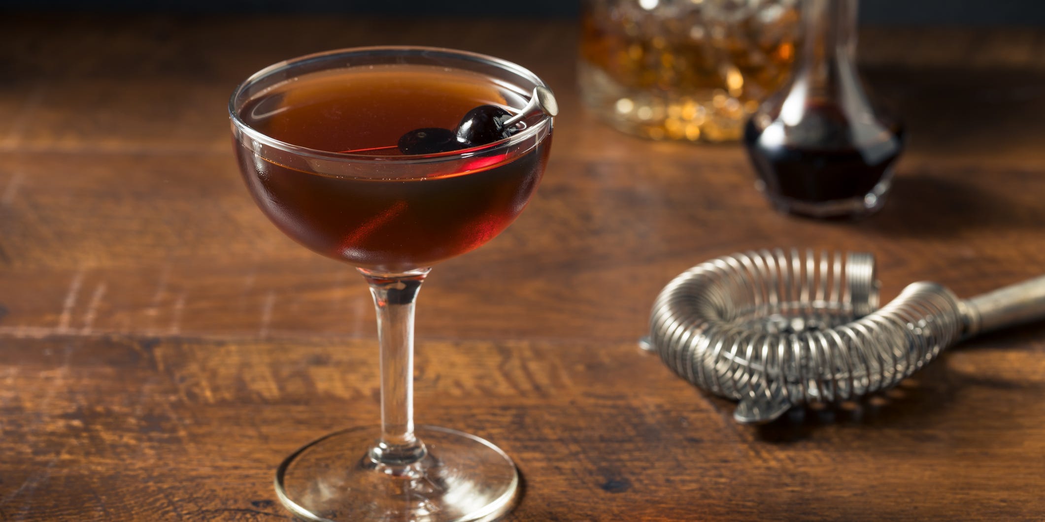 A Manhattan cocktail in a coupe glass garnished with a cherry. A cocktail strainer and a glass decanter of whiskey sit in the background.