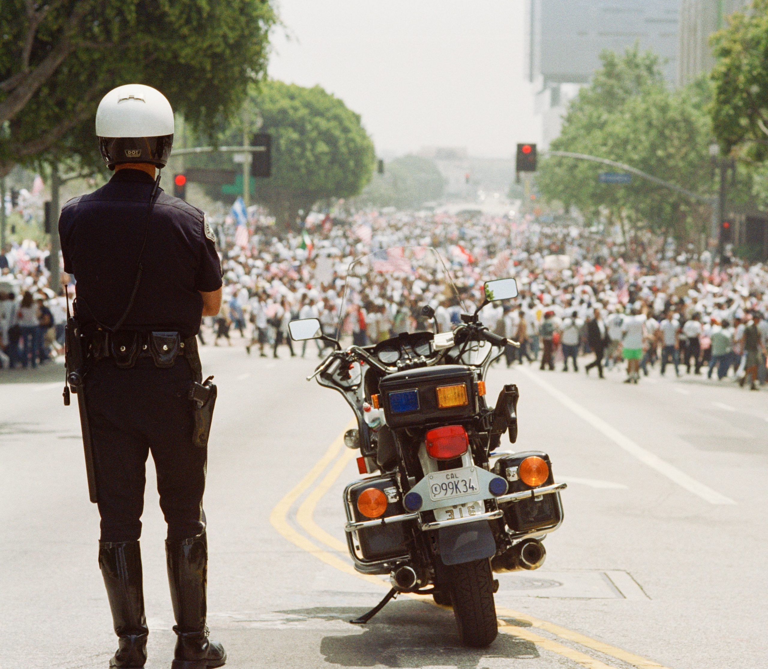 A police officer stands before a crowd of protestors with their motorcycle to the side.