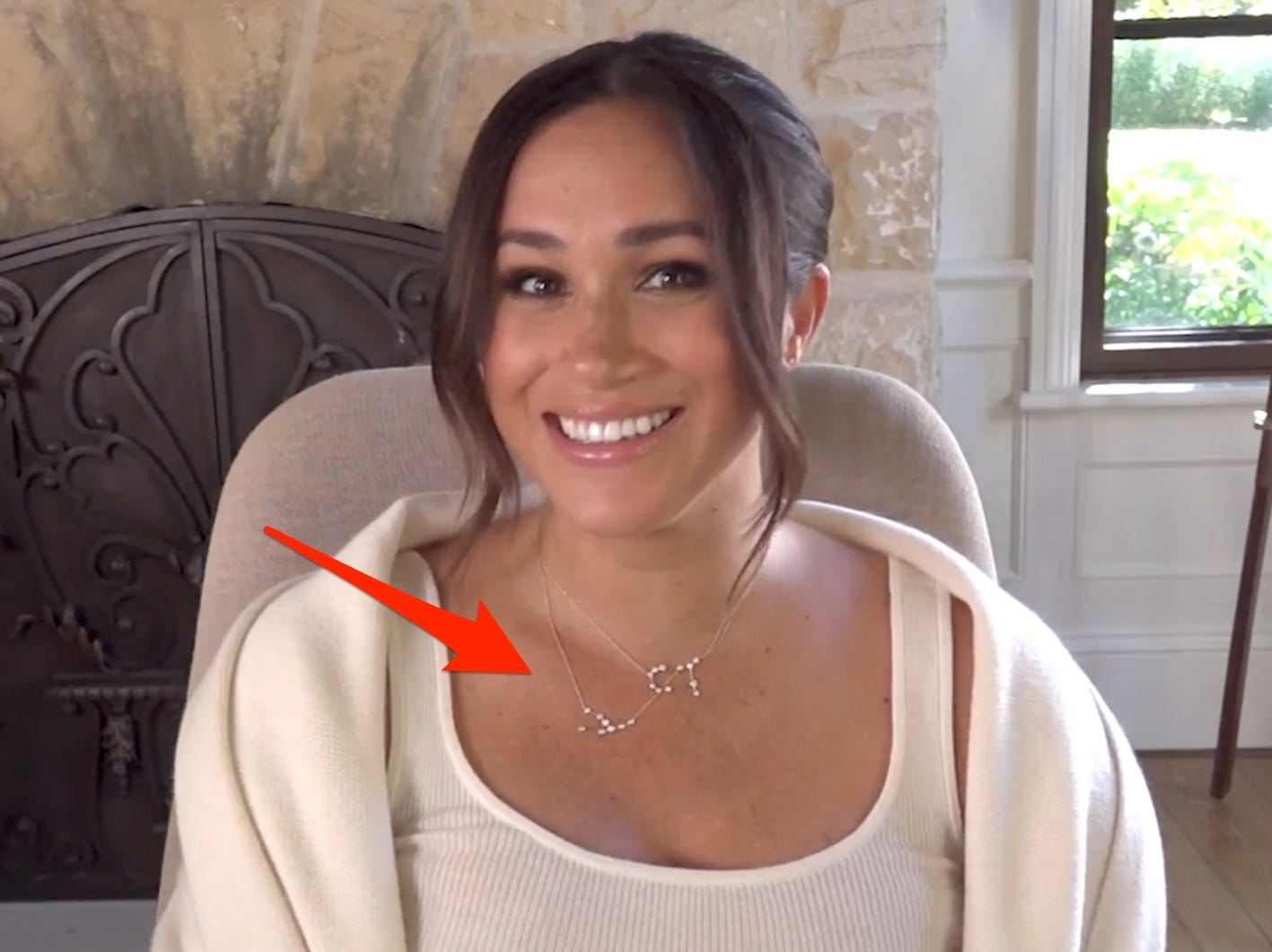 Meghan Markle smiles in a cream sweater and white tank top in front of a fireplace. A red arrow points to her necklaces.