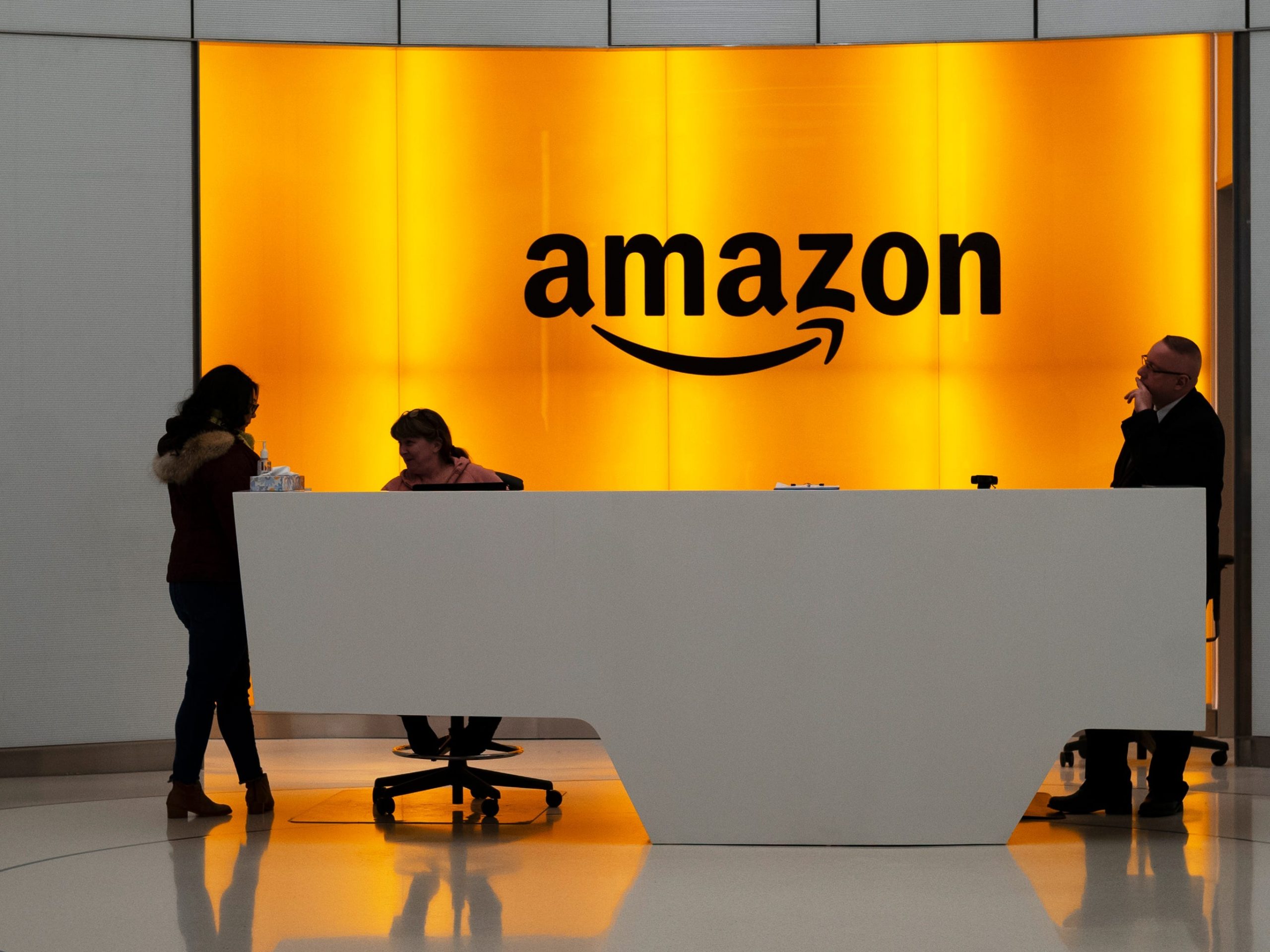 Amazon office front-desk staff stand in front of an orange sign with the Amazon smile logo