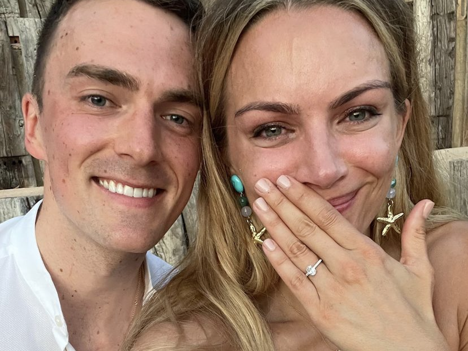 A couple smile for a selfie and the woman raises her left hand to show off her engagement ring.