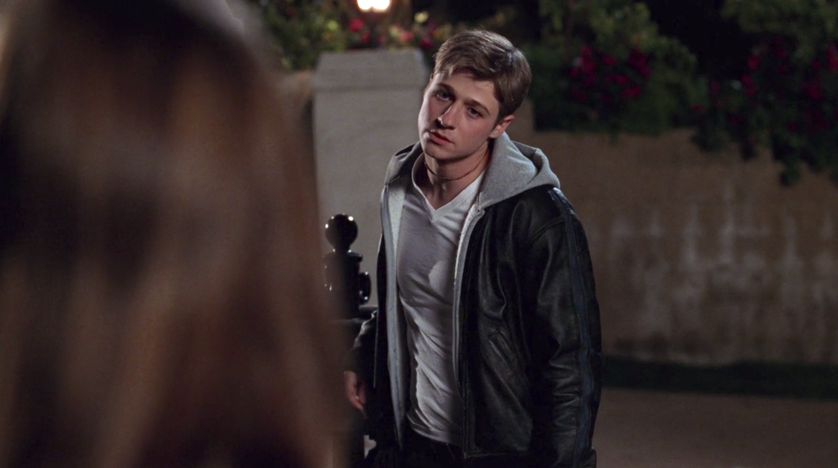 A blonde teenage boy standing outside, wearing a tight white T-shirt under a grey hoodie and black leather jacket.