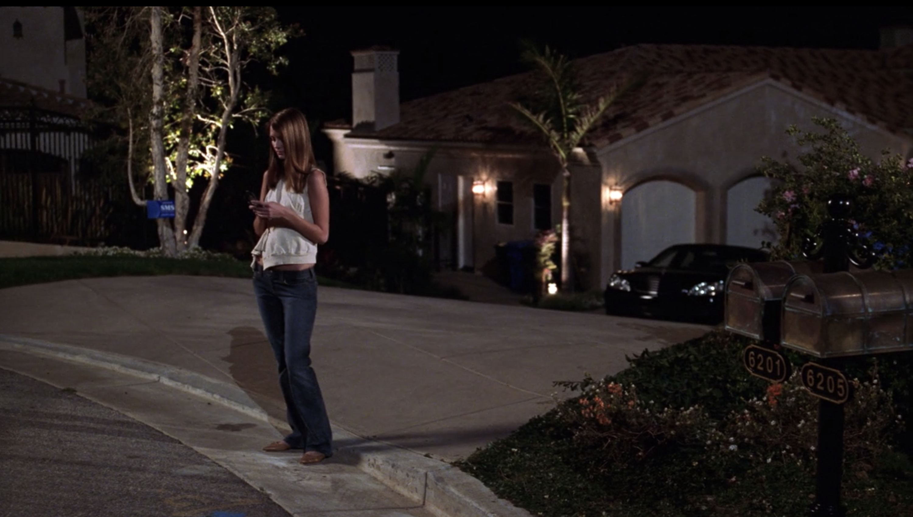 A teenage girl stands on the sidewalk outside a mansion, looking down at her cell phone.