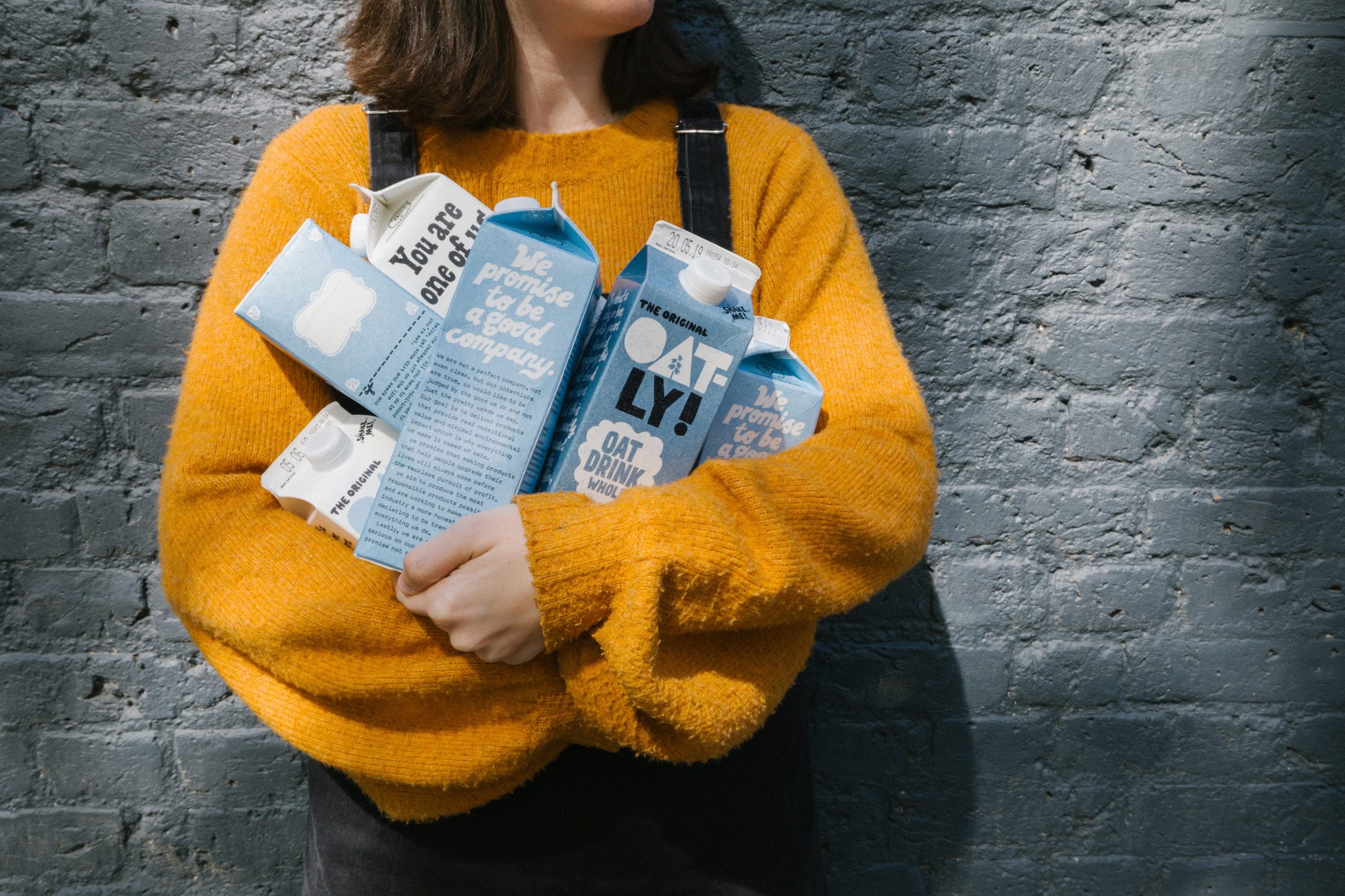 A woman in a yellow sweater holds a half-dozen blue-and-white Oatly cartons