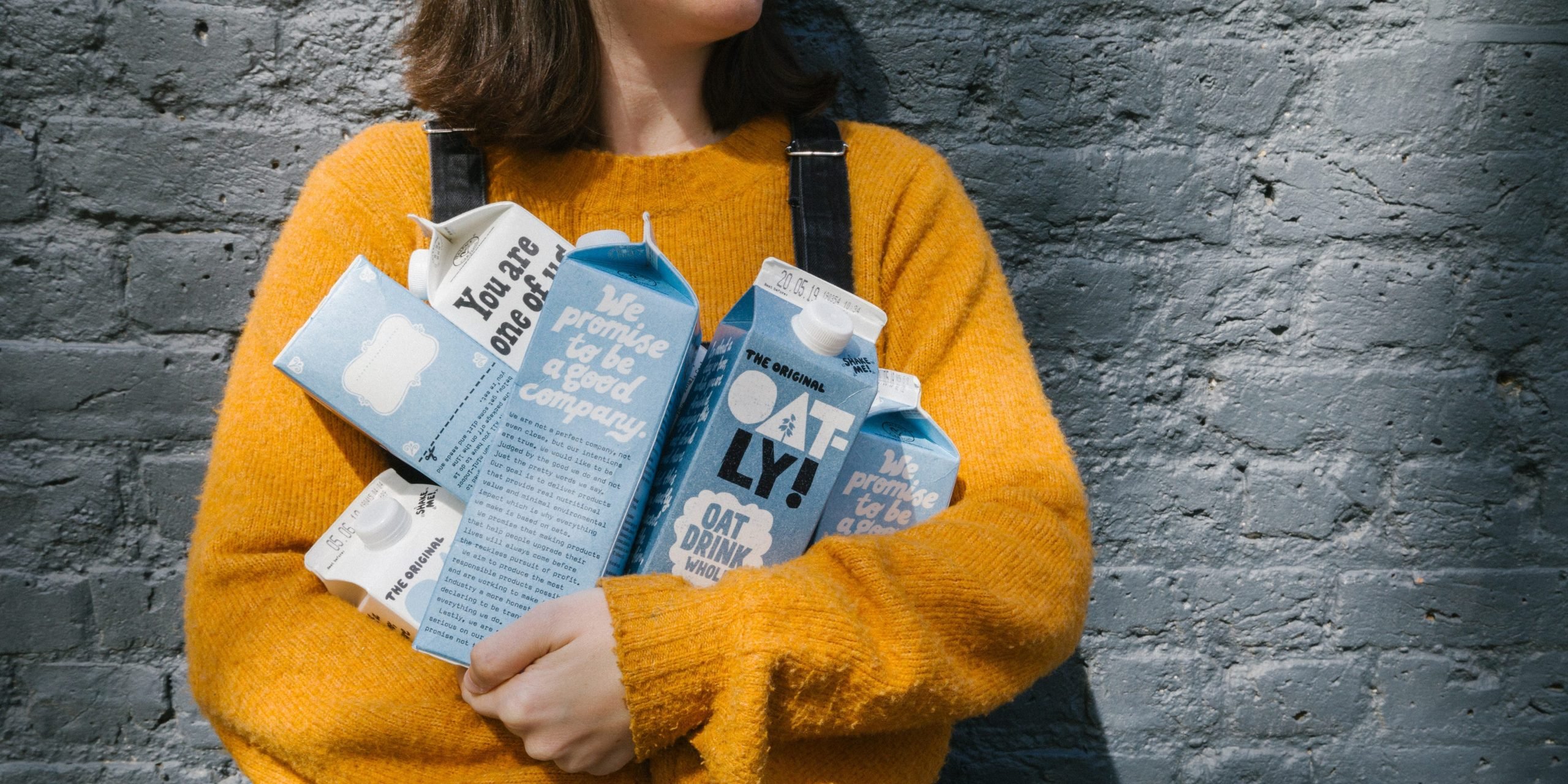 A woman in a yellow sweater holds a half-dozen blue-and-white Oatly cartons