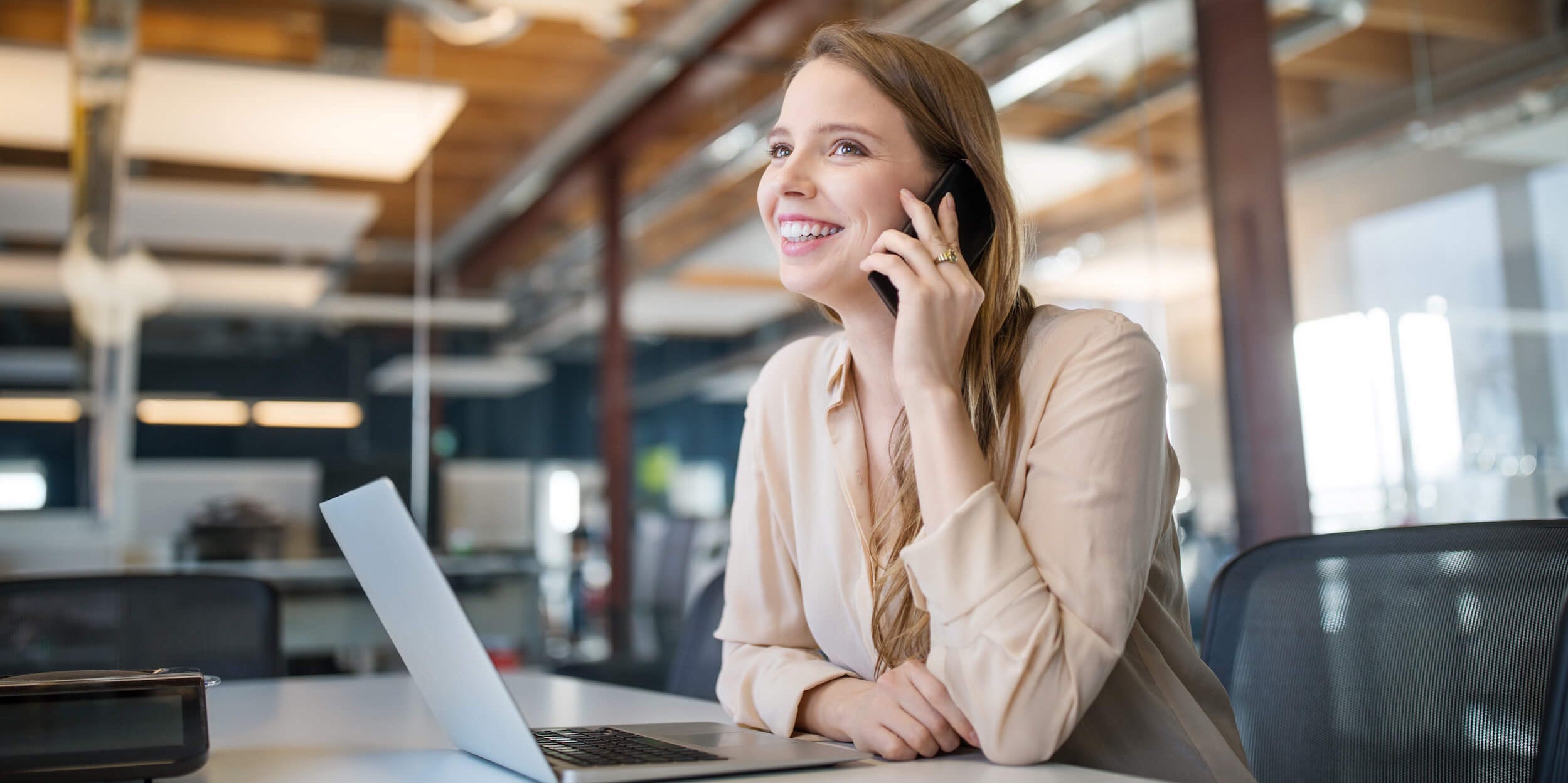 Young businesswoman talking on cell phone while using laptop in an office.