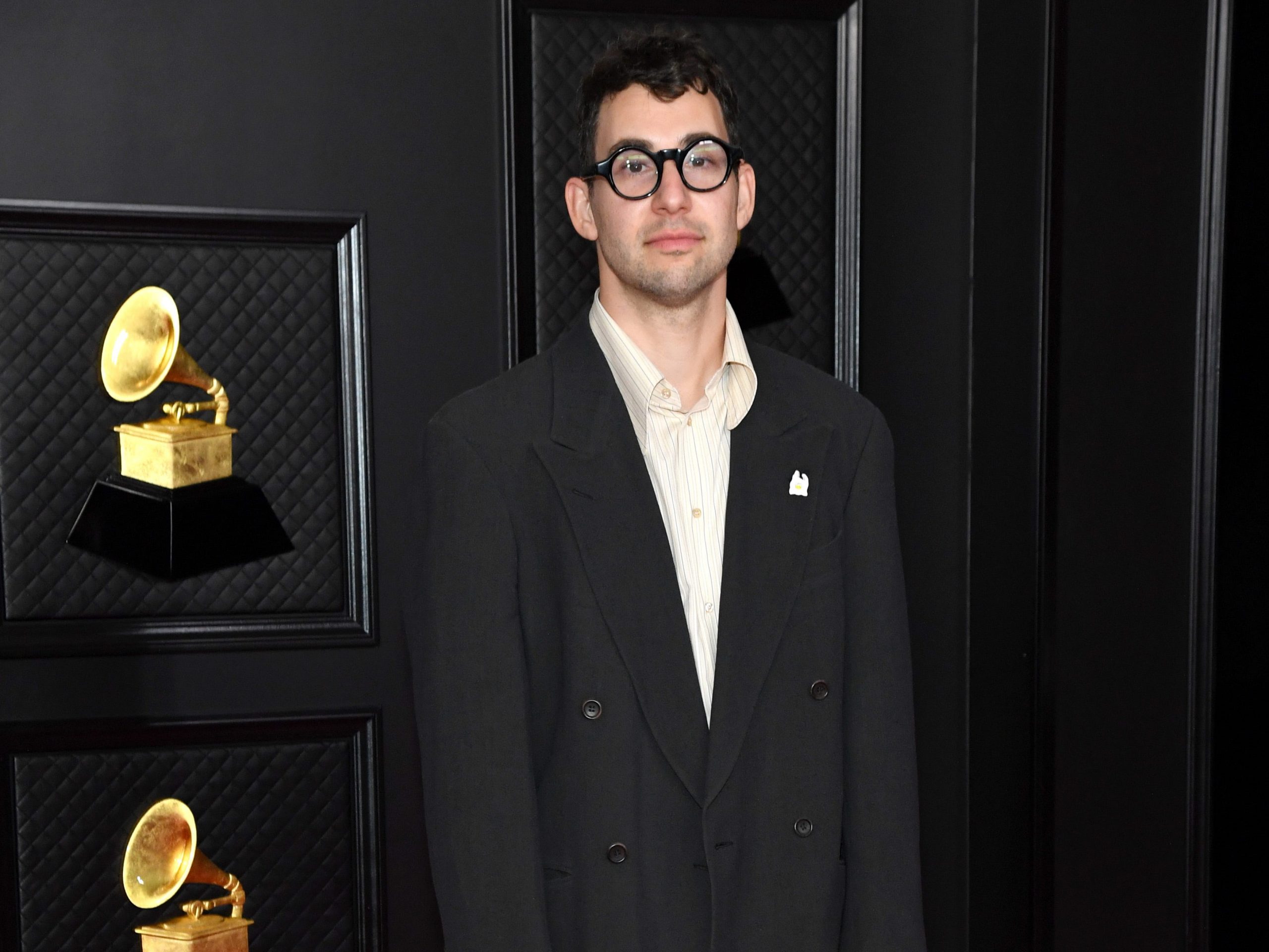 producer jack antonoff standing on a red carpet in a large black suit jacket and brown pants, with the grammys logo behind him. he has a neutral expression on his face