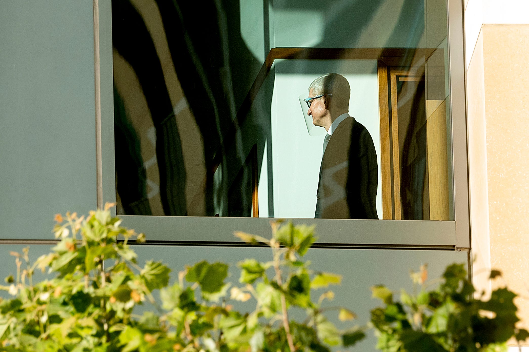 Apple CEO Tim Cook wears a suit and a clear face shield behind a window on a sunny day