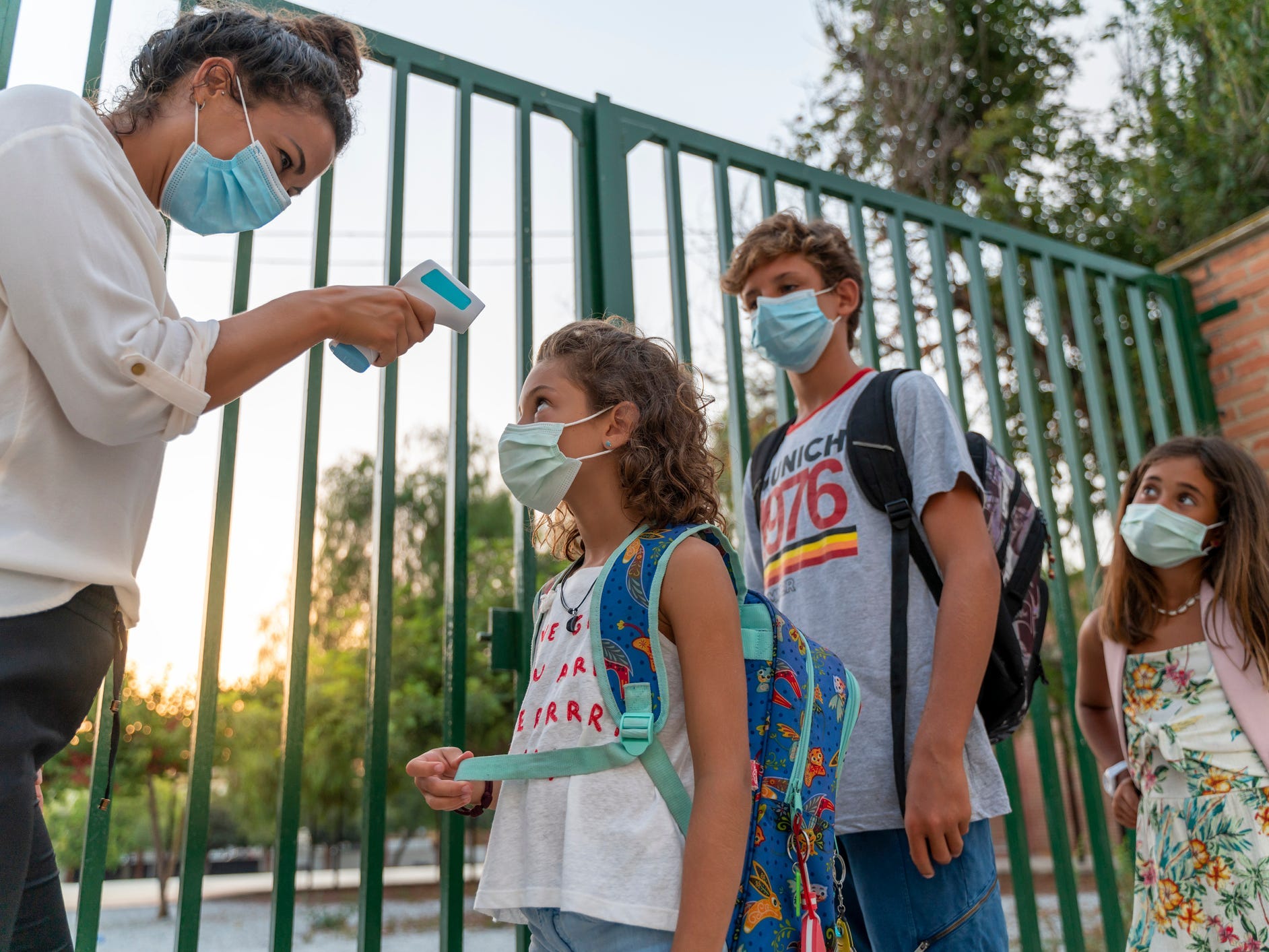 A teacher takes the temperature of pupils lining up outside school wearing face masks.