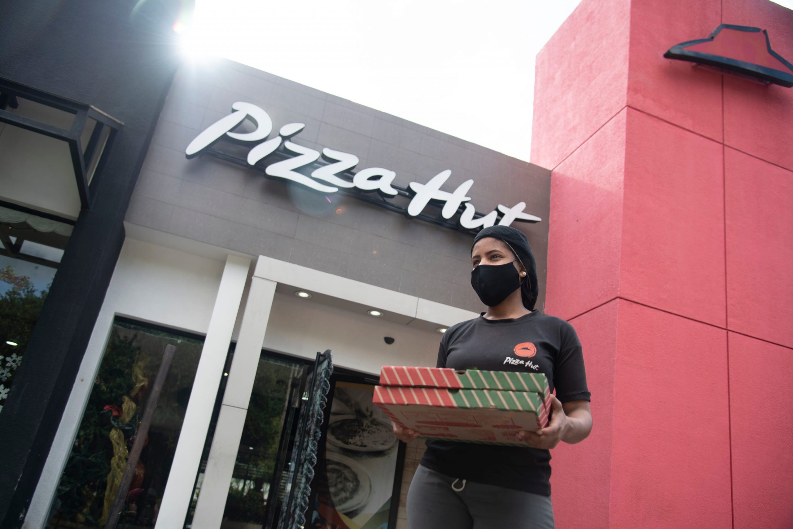 A Pizza Hut employee walks out of a restaurant with a black and red facade carrying two pizza boxes.