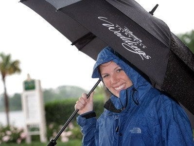 A woman holds at umbrella in a blue raincoat with a palm tree in the background.