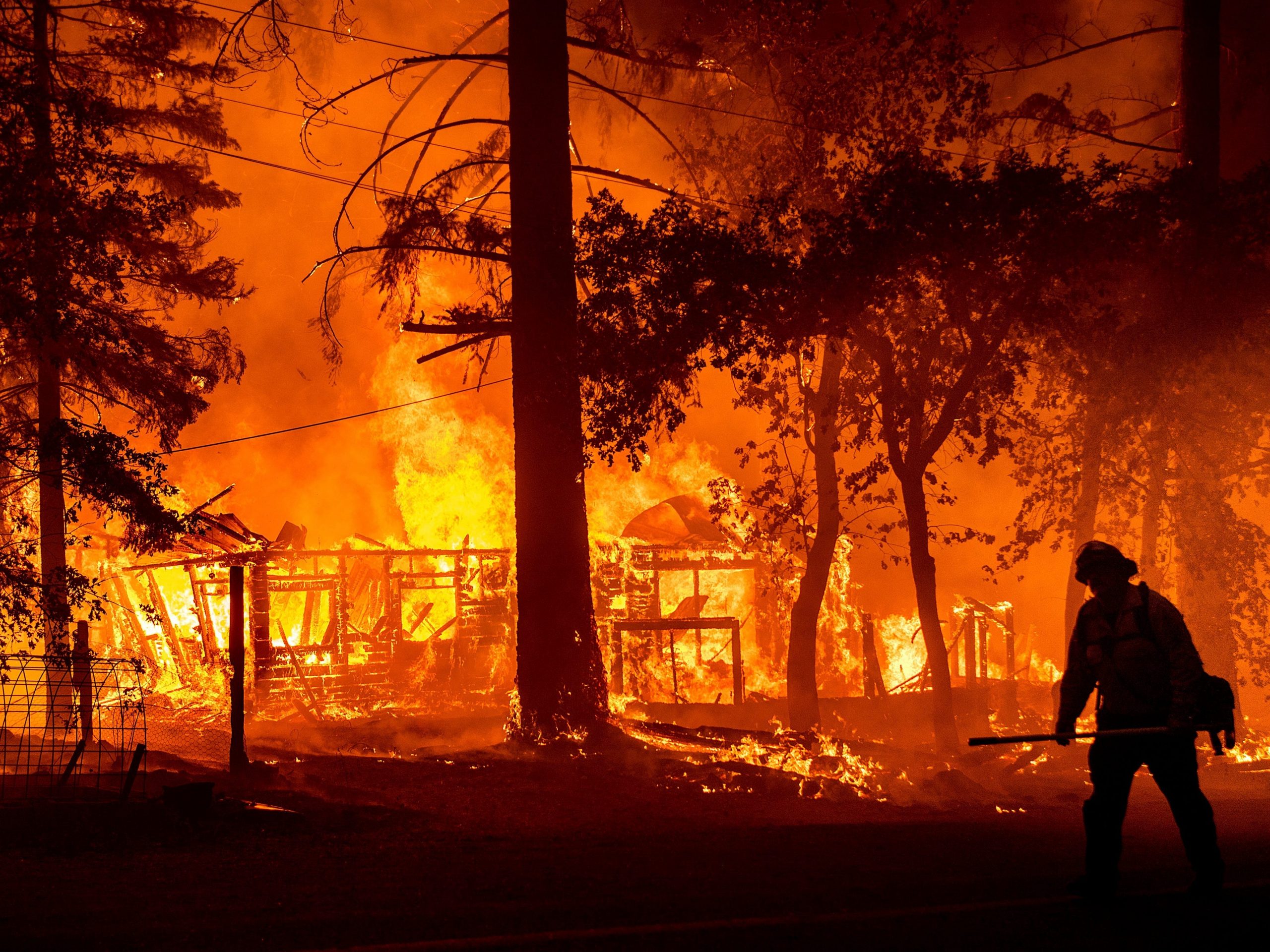 The sillouhette of a firefighter stands in front of a burning home.