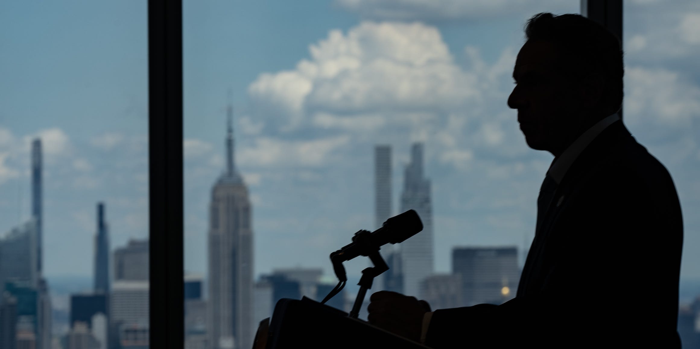 Andrew Cuomo is pictured in silhouette against a New York City skyline view from behind his podium at an event.