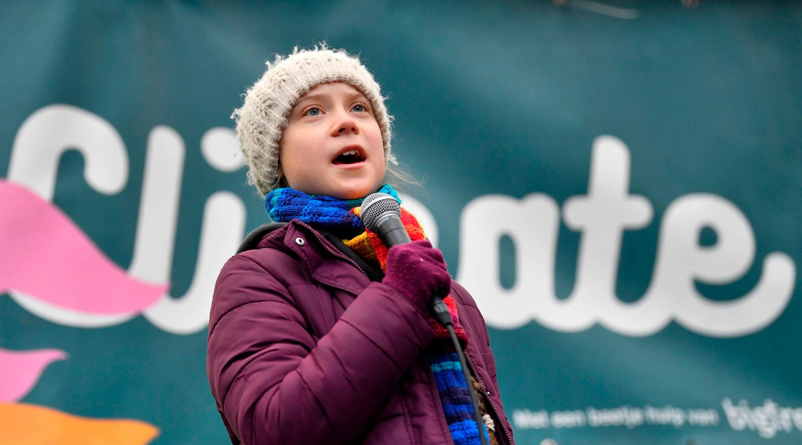 Greta Thunberg, wearing a grey hat and rainbow scarf, holding a microphone while speaking at a protest in Brussels in March, 2020.