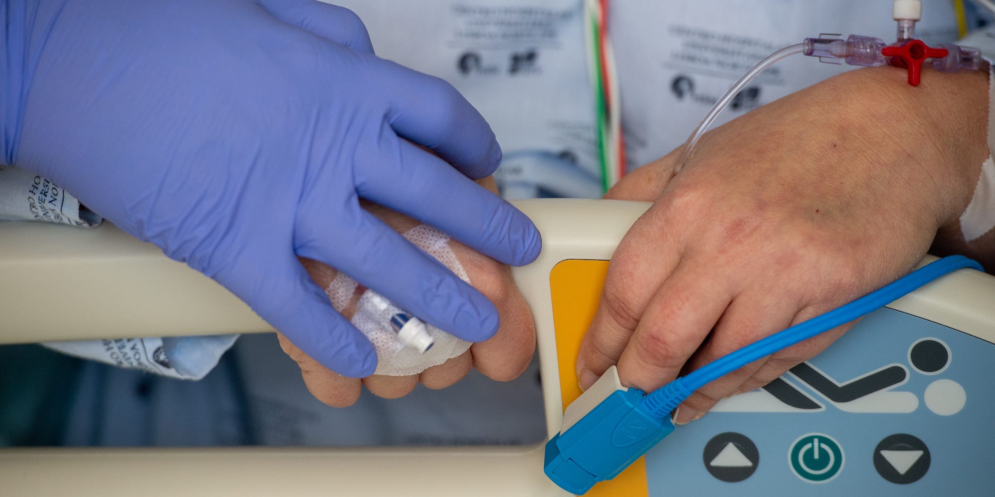 the pictures zooms on the hands of two people holding on to the rail on a hospital bed. One is wearing a blue PPE glove